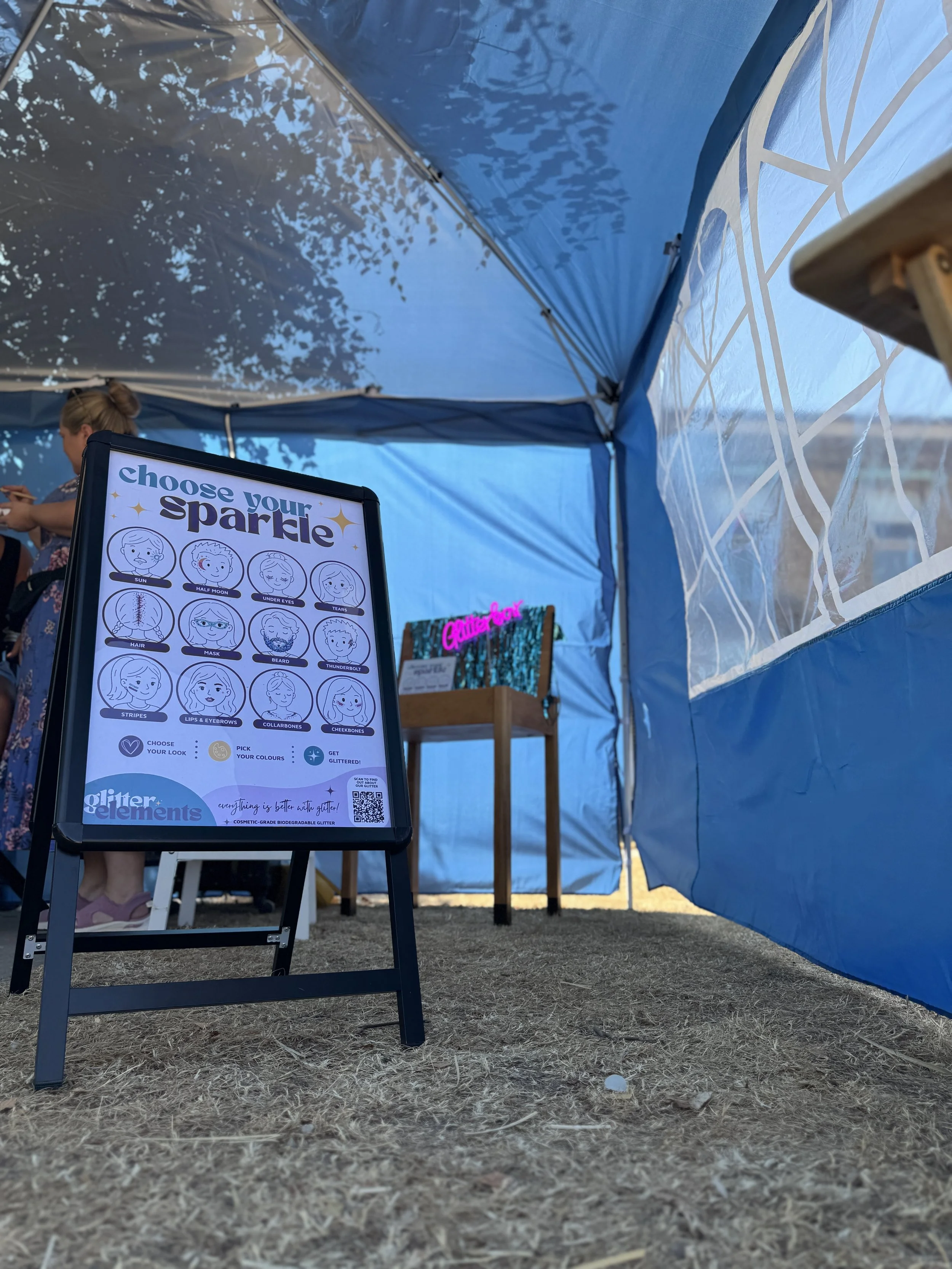 An outdoor event booth with a sign advertising a craft activity called 'Choose Your Sparkle.' The sign shows different face parts and features for decoration, such as eyes, lips, eyebrows, and more. The scene is under a blue canopy tent, with a perso