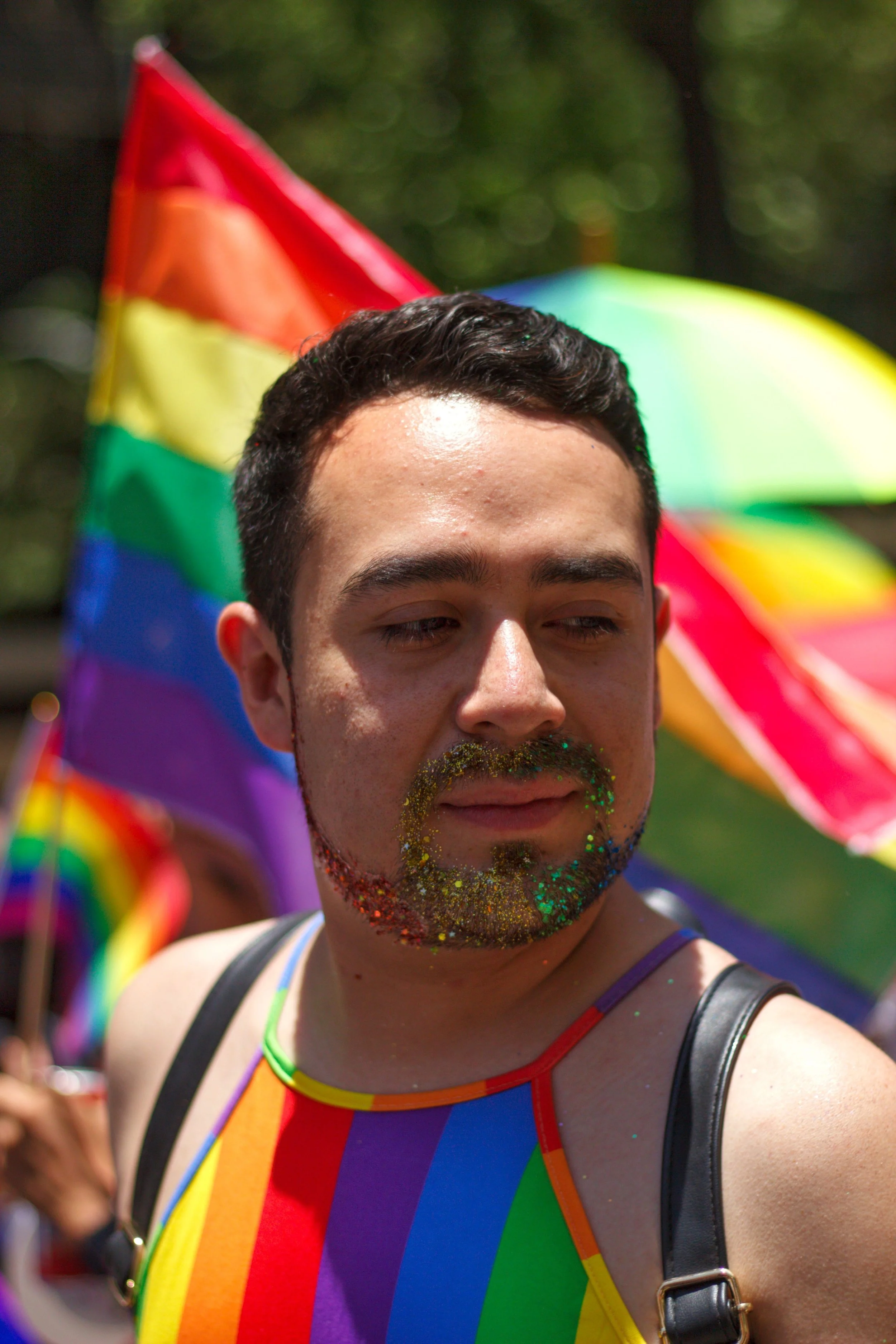 A person with short dark hair and glitter on their beard, wearing a rainbow-colored tank top, participating in a pride event with a rainbow flag and umbrella in the background.