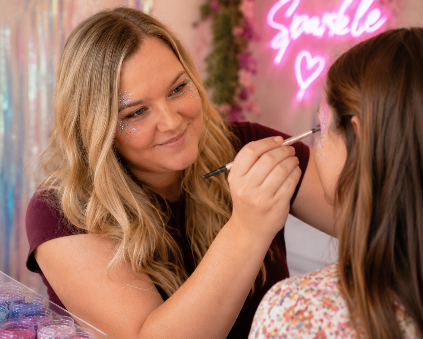 A woman with long blonde hair applying pink and white glitter face paint to a young girl’s face in a decorated room. The woman has face glitter around her eyes and is smiling. Behind them, a neon pink sign reads 'Smile' with a heart, and colorful decorations are visible.