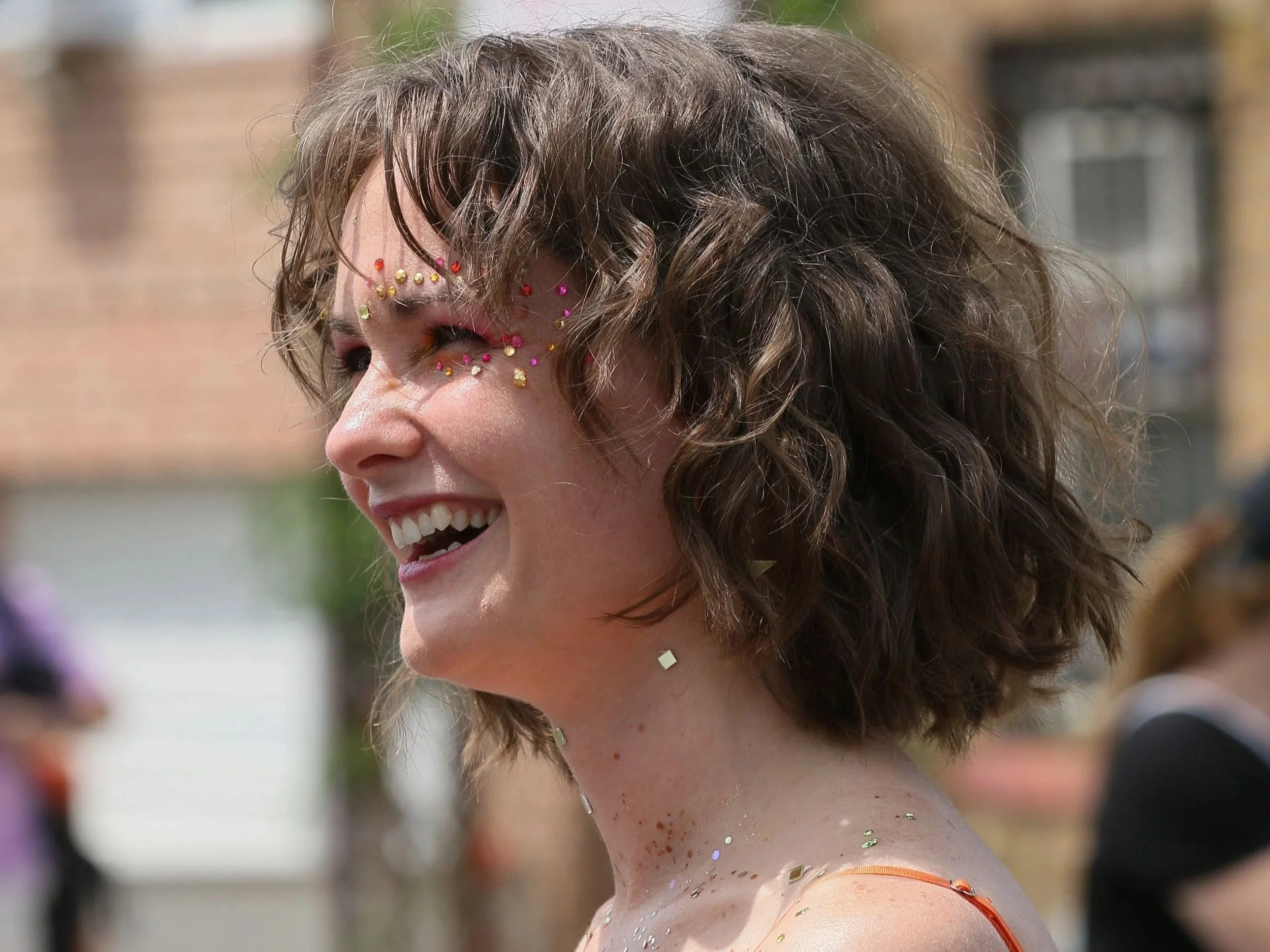 A young woman with curly hair smiling, decorated with colorful rhinestones on her face.