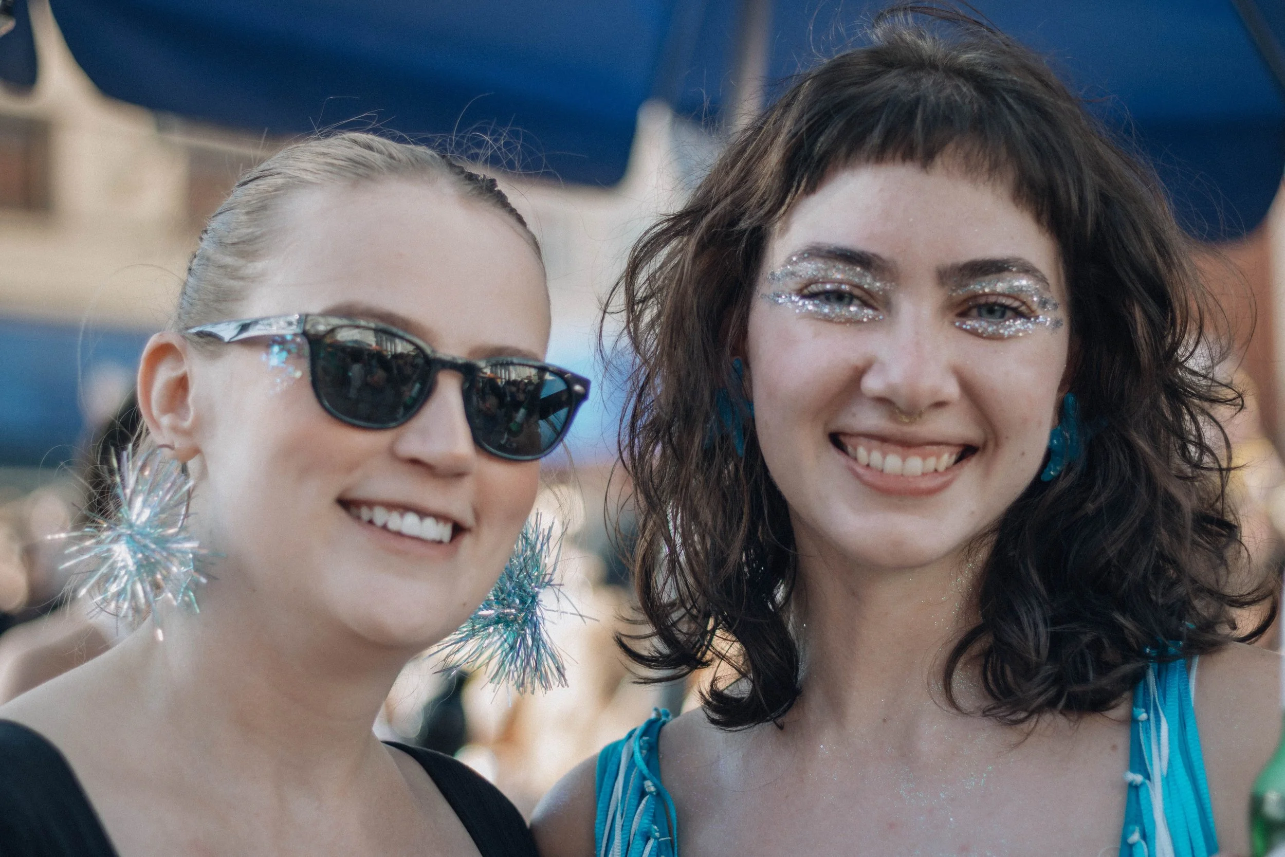 Two young women smiling at a festive celebration, wearing sunglasses and glitter makeup, with blue decorations in the background.