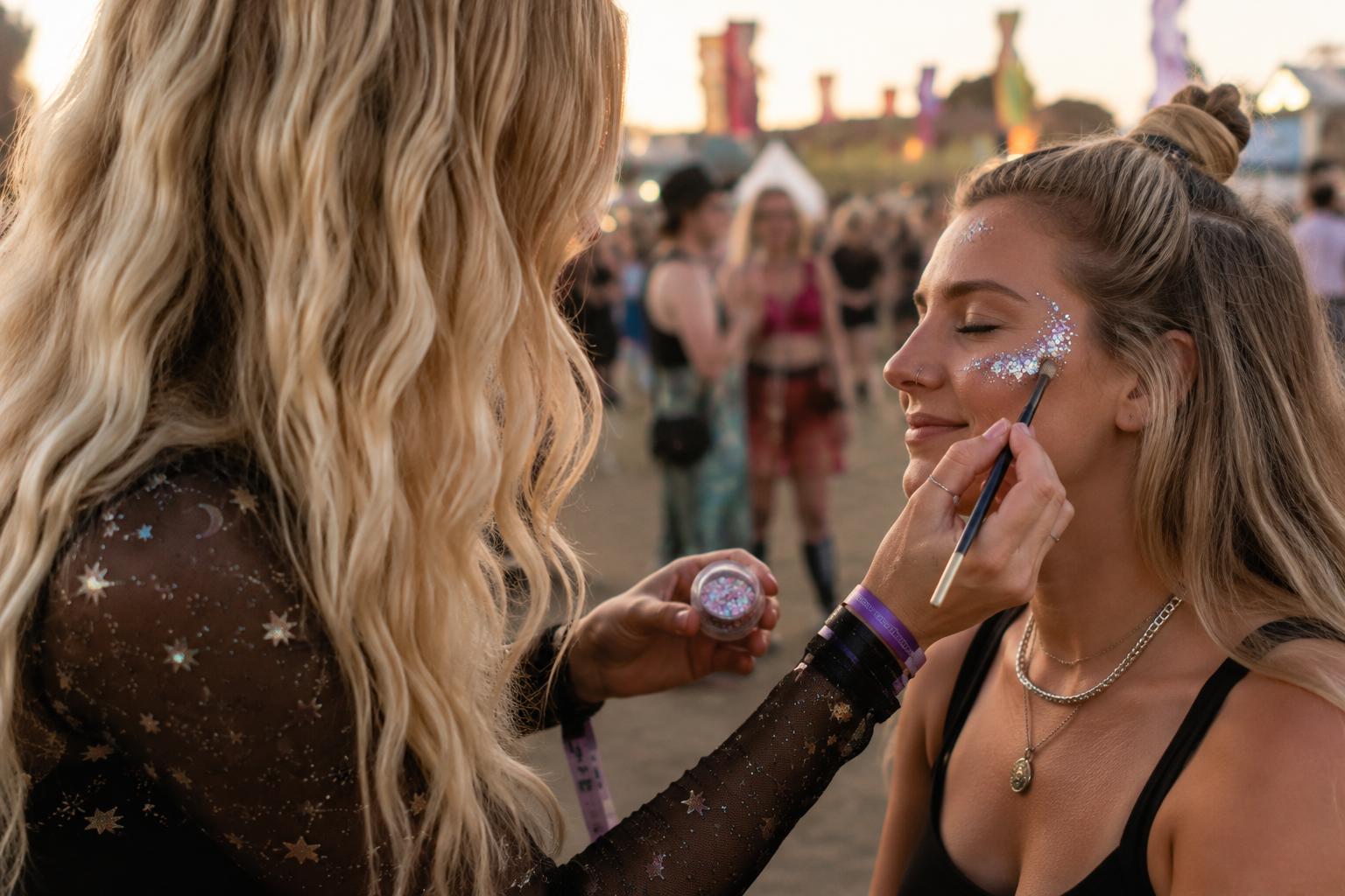 A woman with glitter being applied to her face by another woman at an outdoor event during sunset.