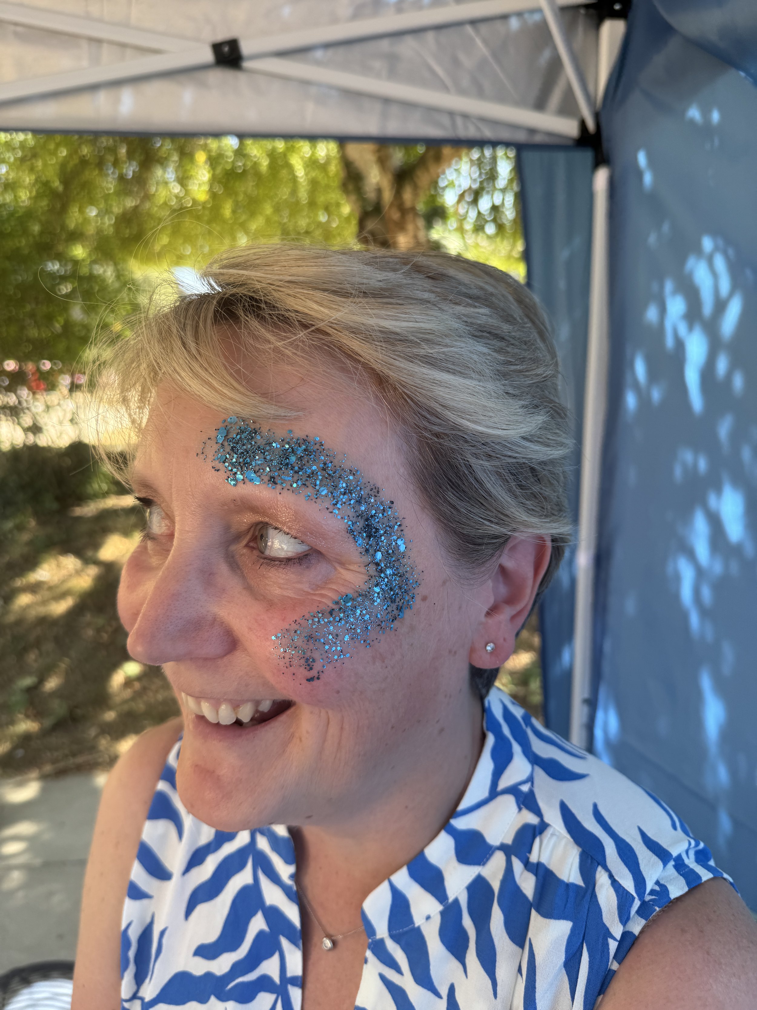 Close-up of a woman with short blonde hair smiling outdoors, wearing blue glitter makeup around her eye, a blue and white patterned top, a small necklace, and small earrings. There is greenery and a tent in the background.