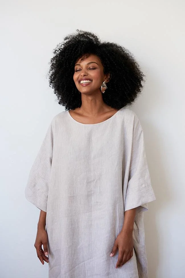 A woman with curly black hair smiling and wearing a white, loose-fitting dress with wide sleeves.