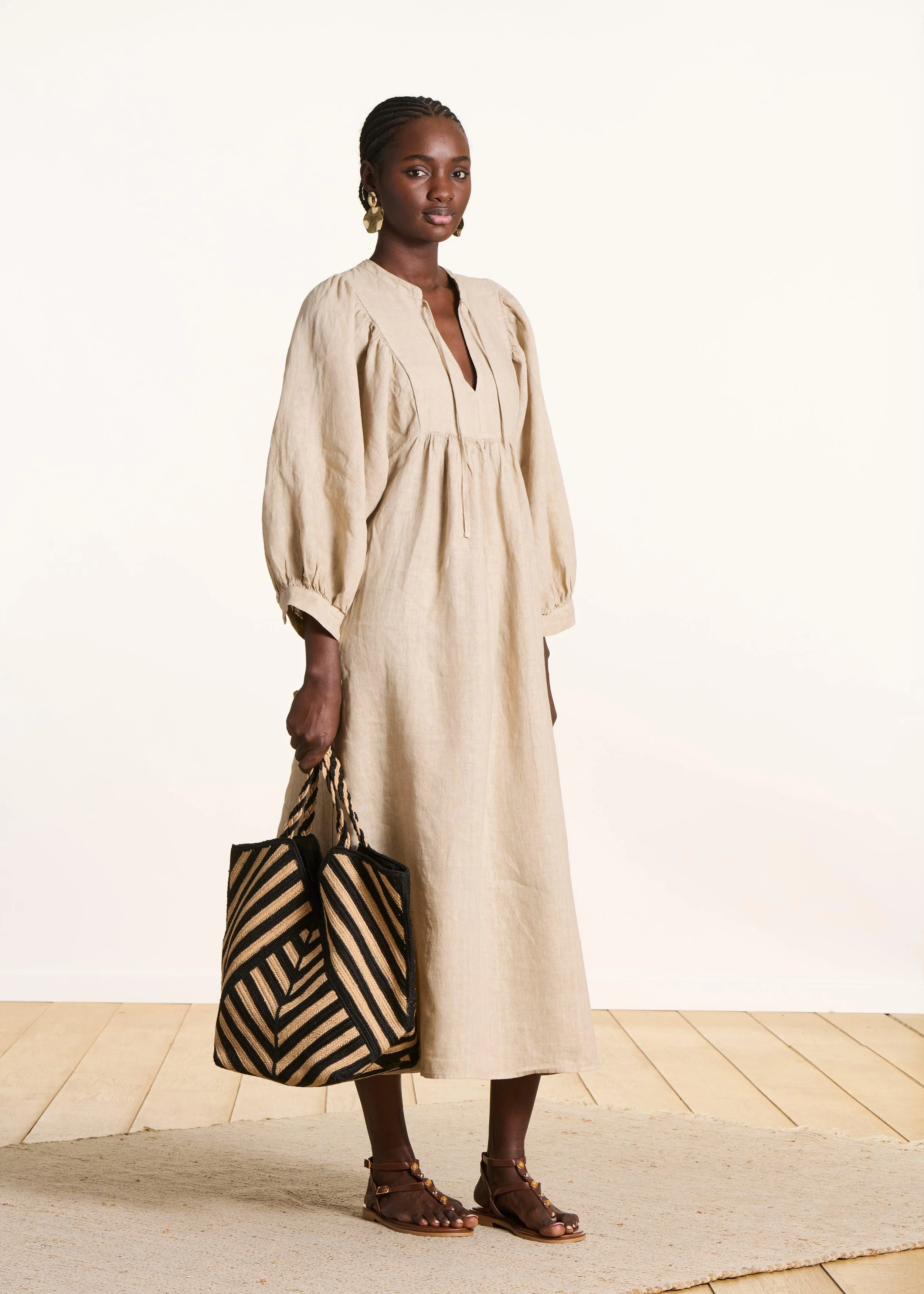 A woman with dark skin and braided hair stands on a beige rug in front of a plain off-white wall. She is wearing a loose beige dress with puffed sleeves and a V-neck, gold earrings, and brown gladiator sandals. She holds a black and beige striped tote bag.