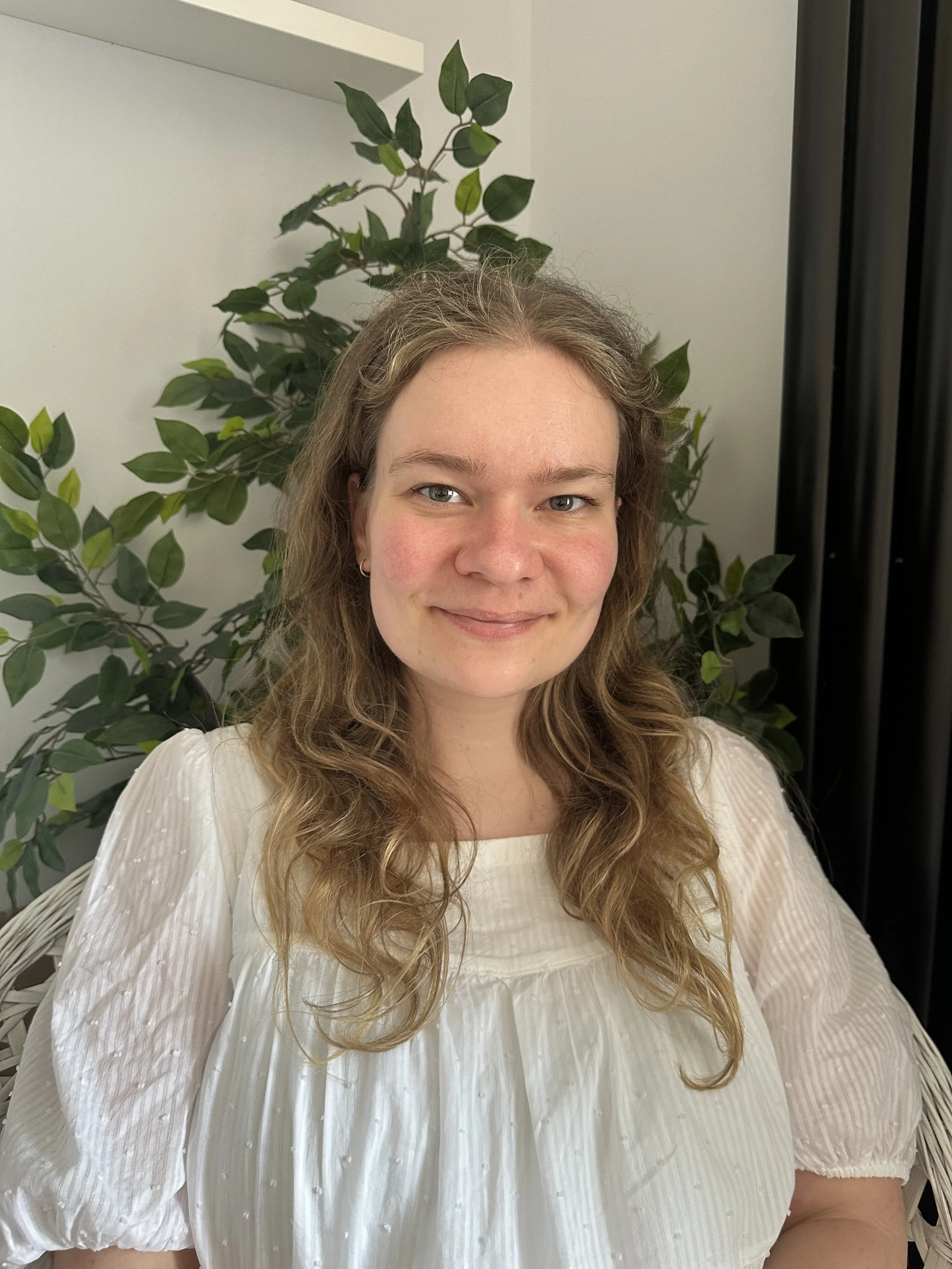 Jess, a woman with long, wavy, light brown hair and fair skin, smiling, wearing a white, textured, semi-sheer blouse with puffed sleeves, sitting in front of a green leafy plant and a white wall. She is a a fully qualified therapist and counsellor.