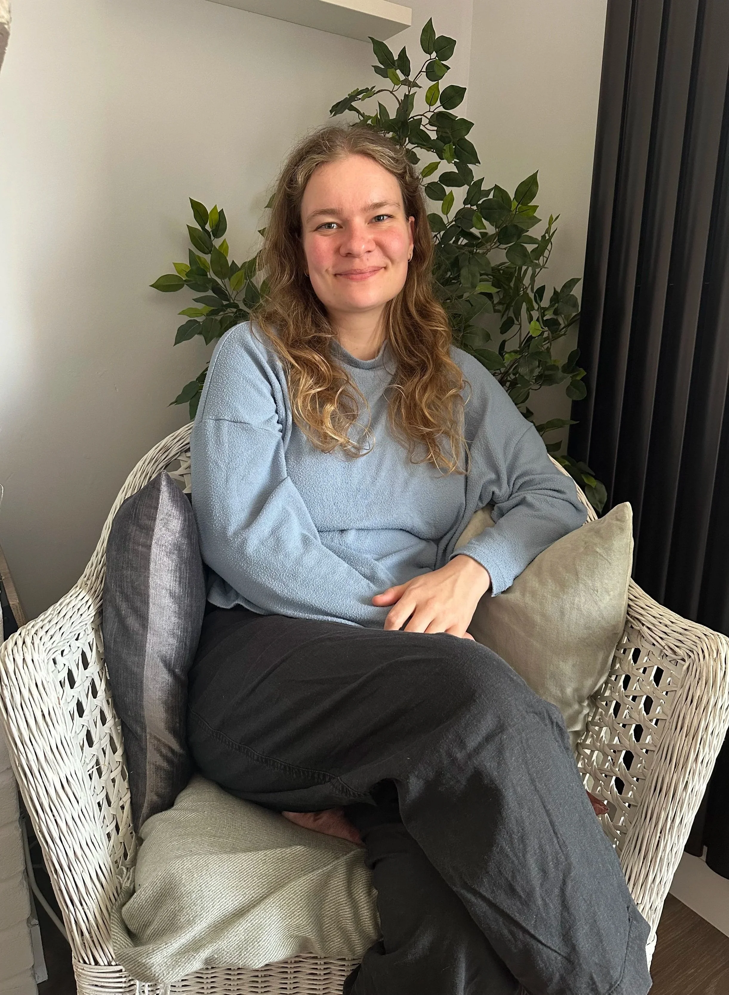 Jess, a fully qualified therapist and psychotherapeutic counsellor, is a young woman with long, wavy brown hair.  She is smiling, with a large leafy plant behind her and a black partition to her right.