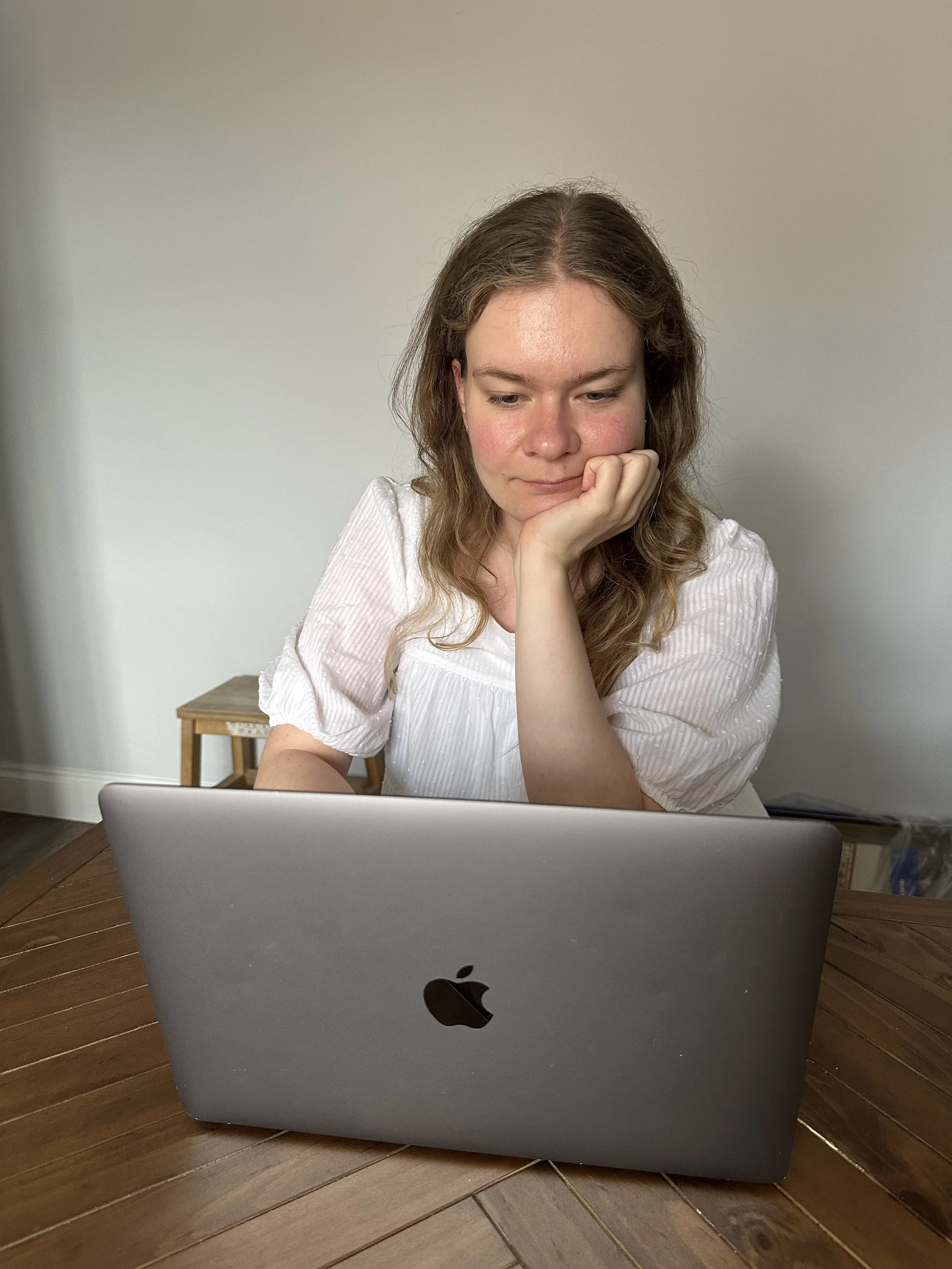 Jess, a woman with wavy brown hair looking down at a laptop, sitting at a wooden table in a room with light-colored walls. She is a fully qualified therapist and psychotherapeutic counsellor, and is at work.