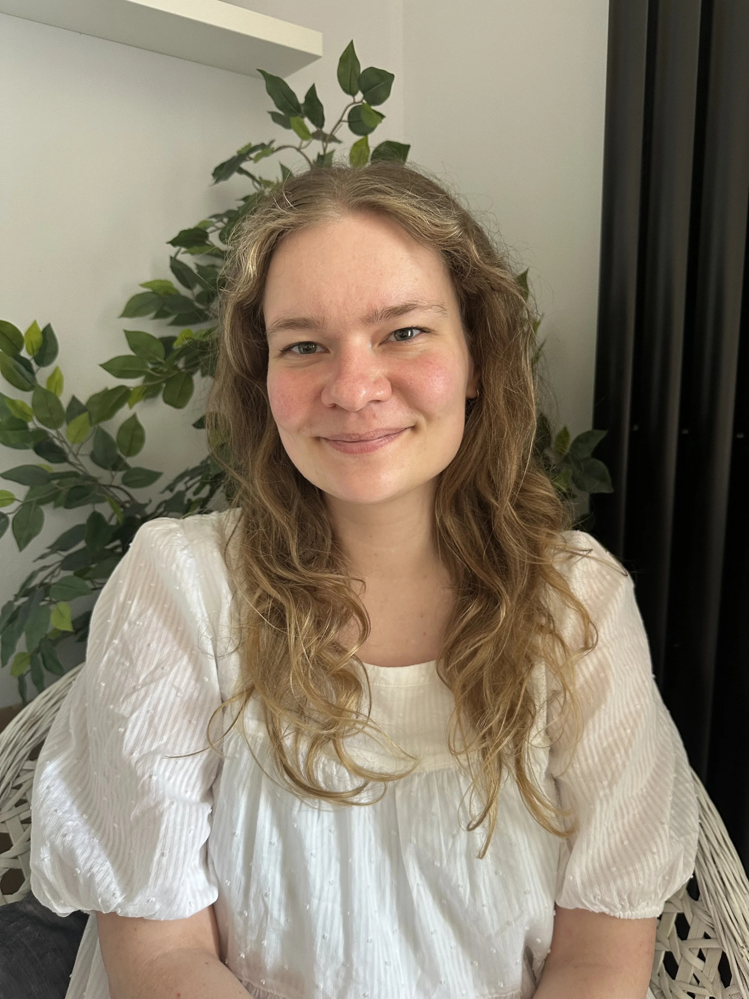 Jess, a woman with long wavy hair smiling, wearing a white pleated blouse, sitting in front of a green leafy plant and a white wall. She is a fully qualified therapist and psychotherapeutic counsellor.