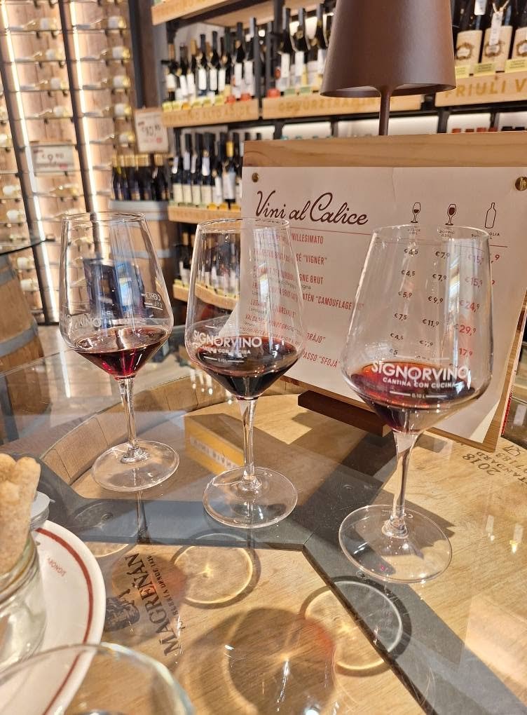 Three glasses of red wine on a wooden table with a wine shop in the background and a menu stand.