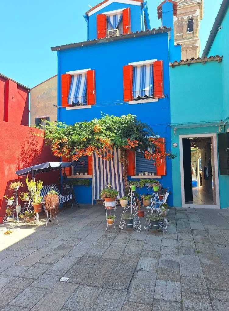Colorful house with blue walls and red window shutters, surrounded by potted plants and outdoor furniture on a stone patio.