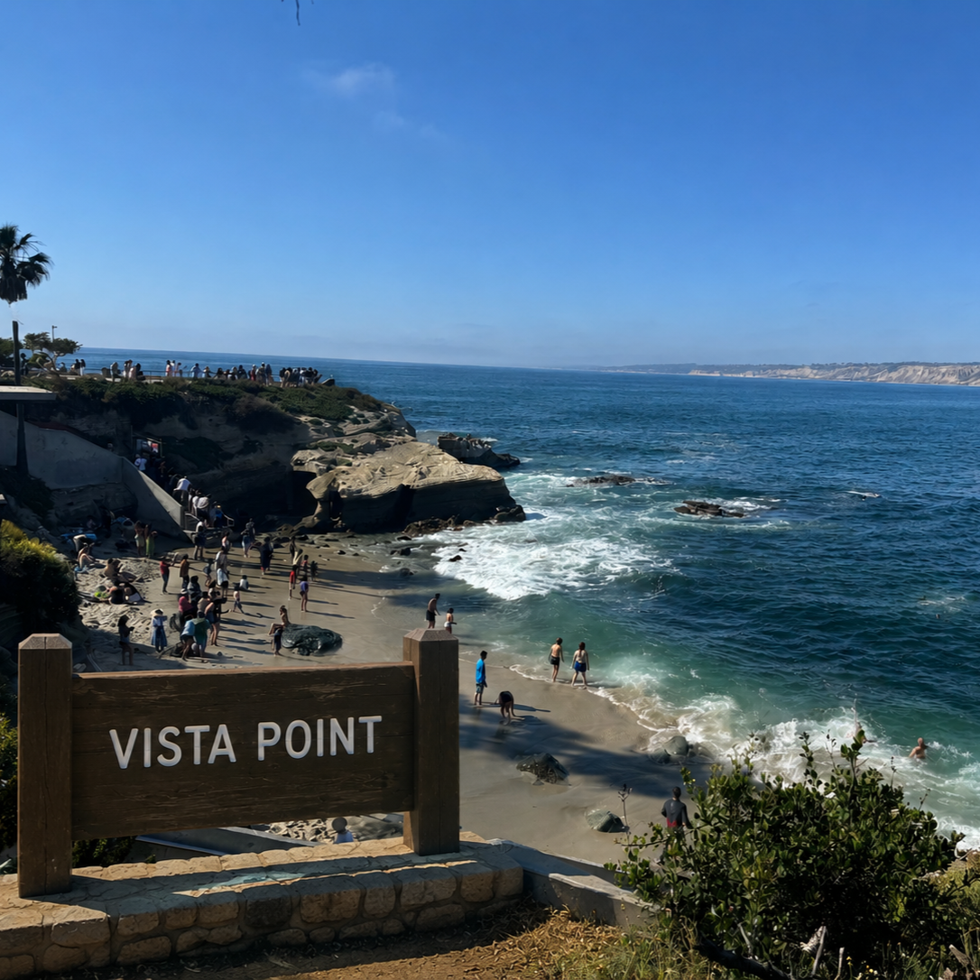 View of Vista Point overlooking a beach with many people, rocky cliffs, and the ocean on a clear day with blue sky.