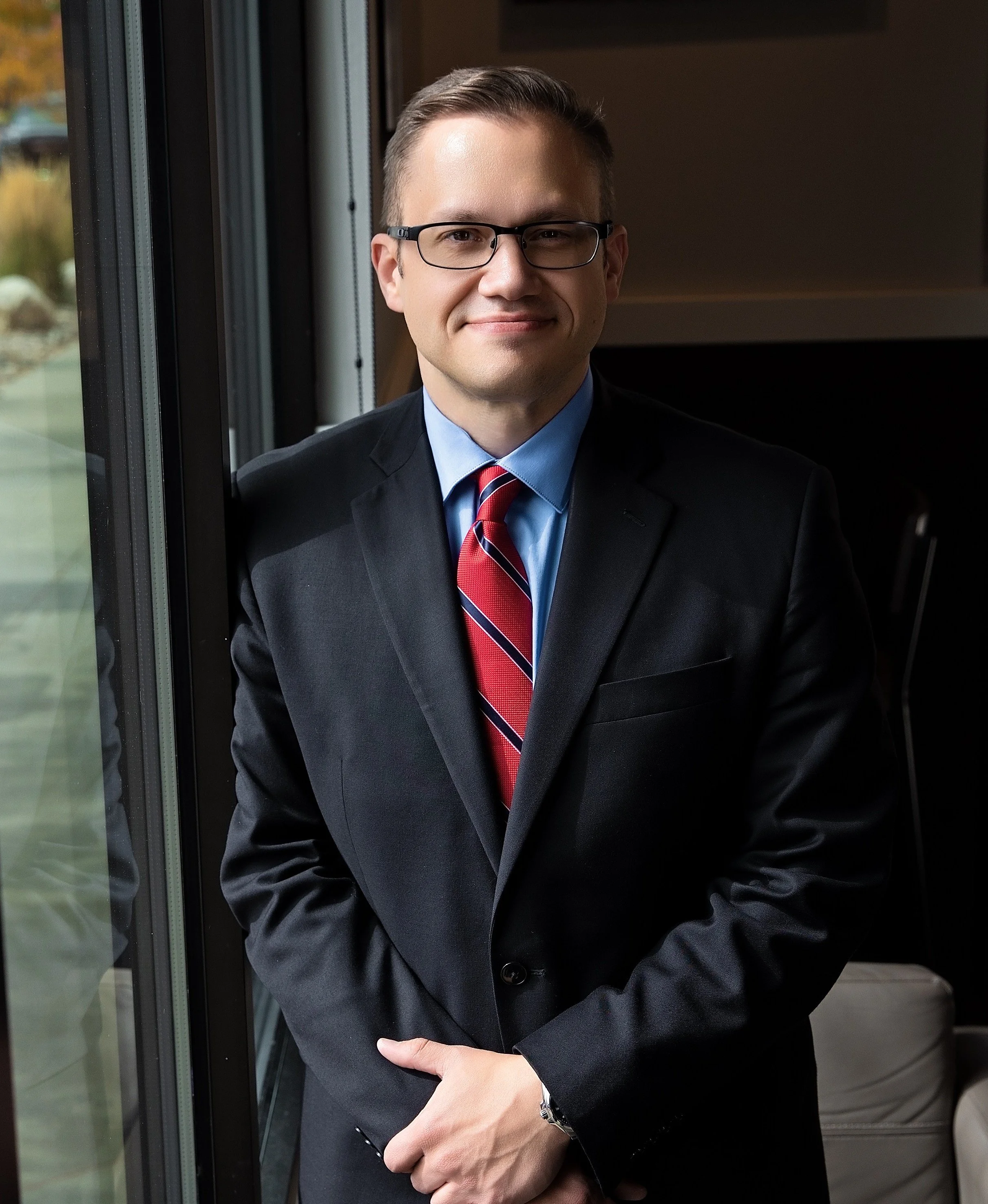 Kyle Ferris in a dark suit, light blue shirt, and red striped tie, wearing glasses, smiling and standing near a window with a blurred outdoor background.