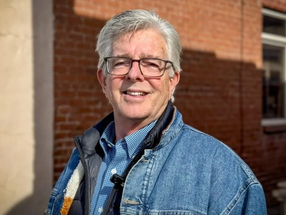 A smiling middle-aged man with glasses and gray hair, wearing a blue checkered shirt and denim jacket, standing outdoors in front of a brick building.