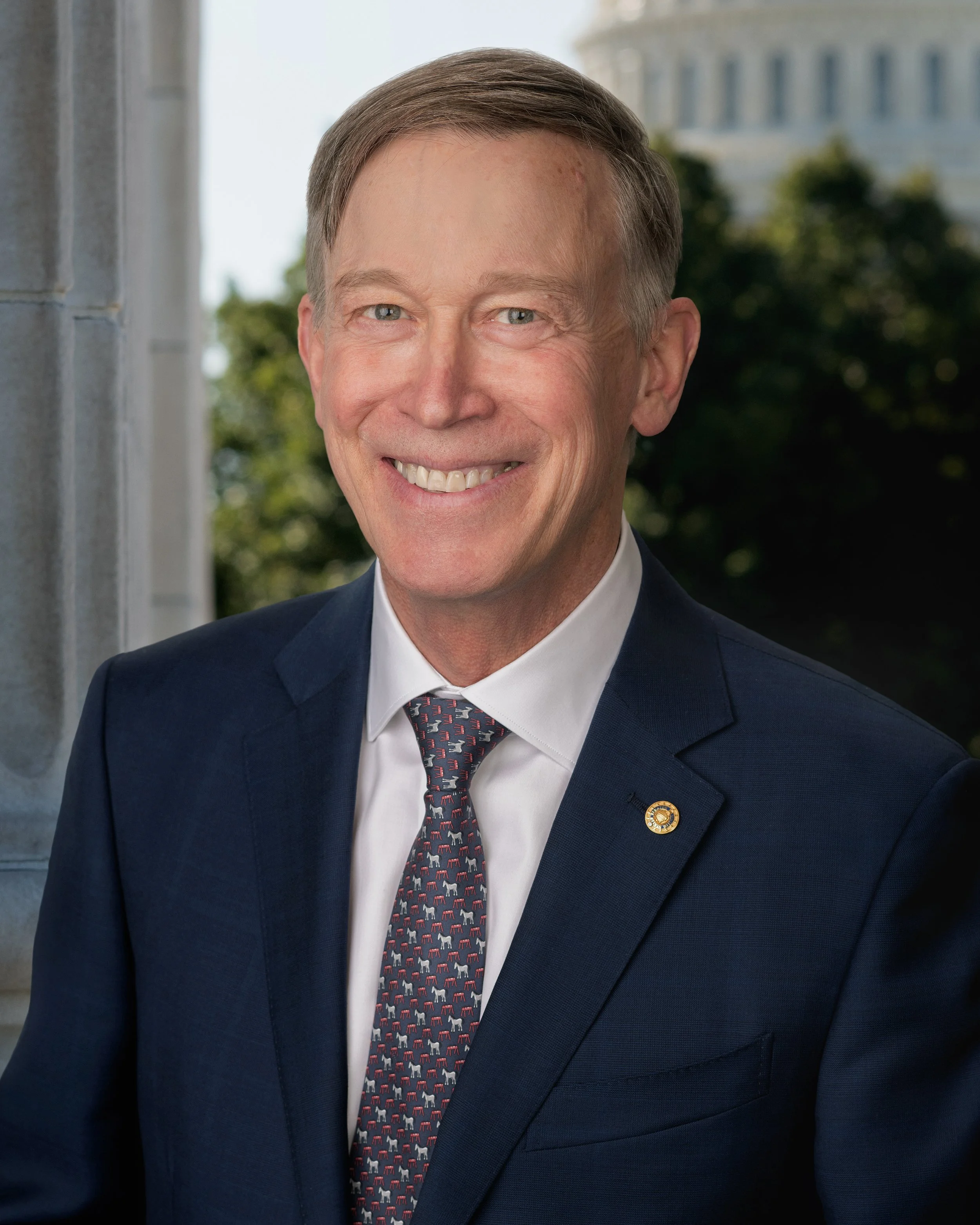 A middle-aged man wearing a navy blue suit, white shirt, and a tie with a pattern of red, white, and blue horses, smiling outdoors with the U.S. Capitol building in the background.
