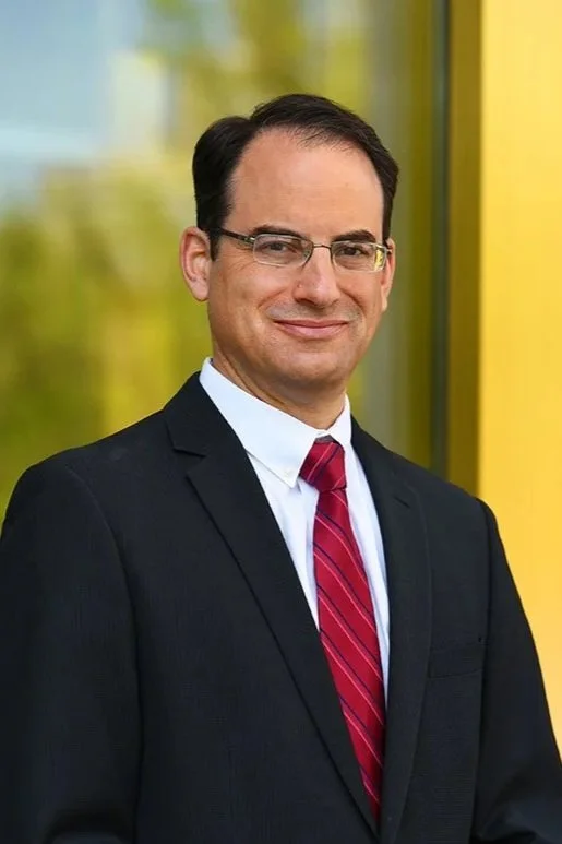 A man in a black suit, white dress shirt, and red striped tie, wearing glasses, standing outdoors in front of a colorful building with yellow and green panels.