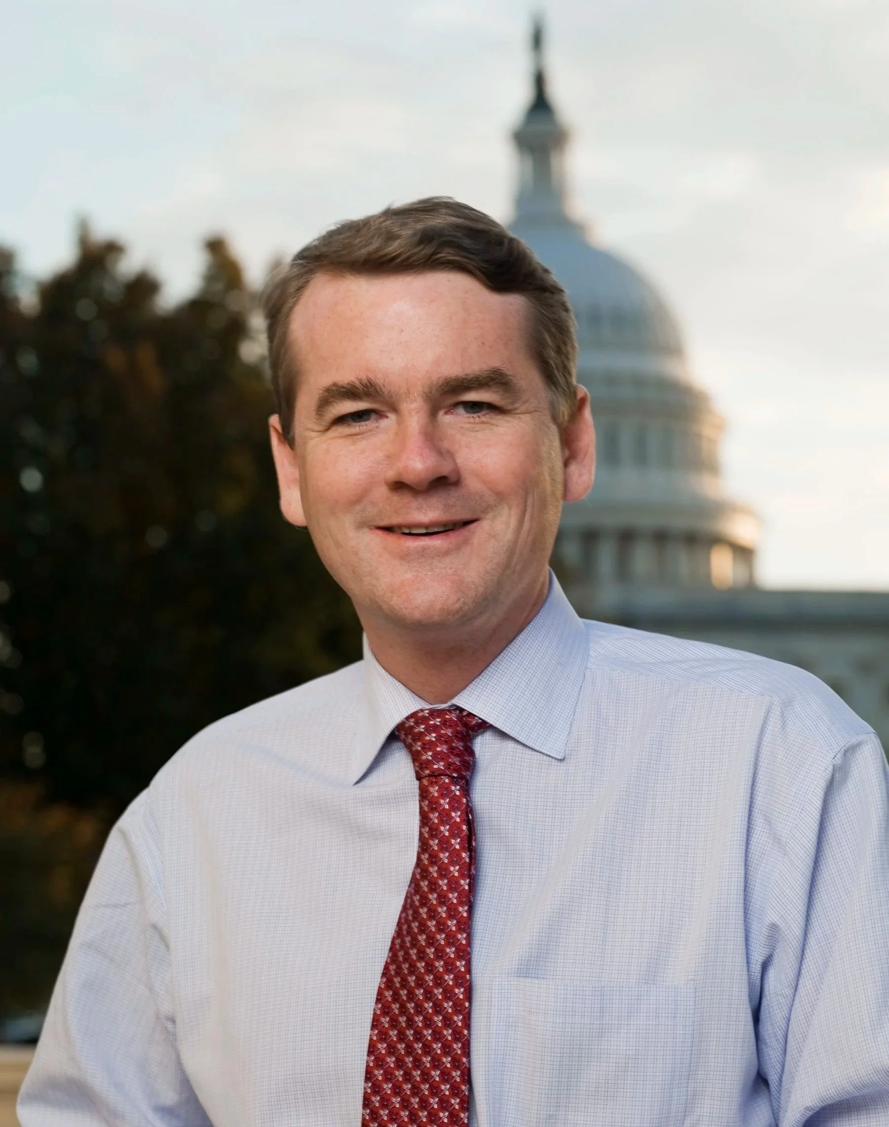 A man in a blue dress shirt and red patterned tie, standing outdoors with the United States Capitol building in the background.
