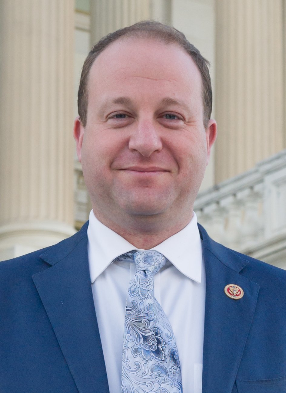 A smiling man in a suit and patterned tie, standing in front of columns and architecture likely in a government building.
