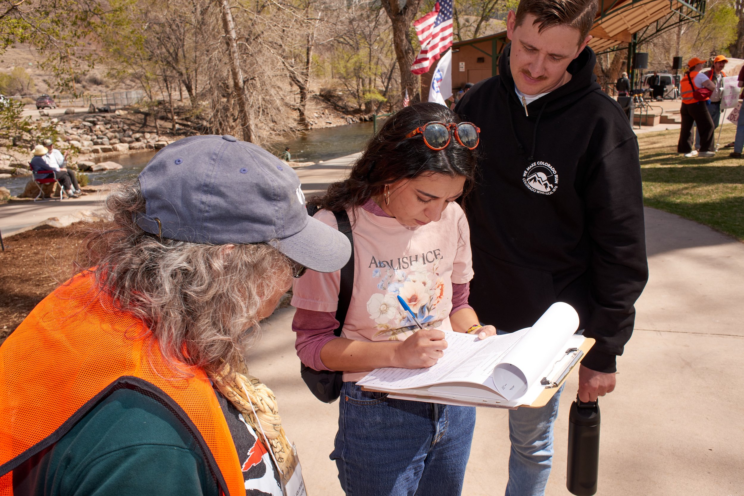 People gathered outdoors by a river, some sitting on benches and others writing or using devices, with trees and flags in the background.