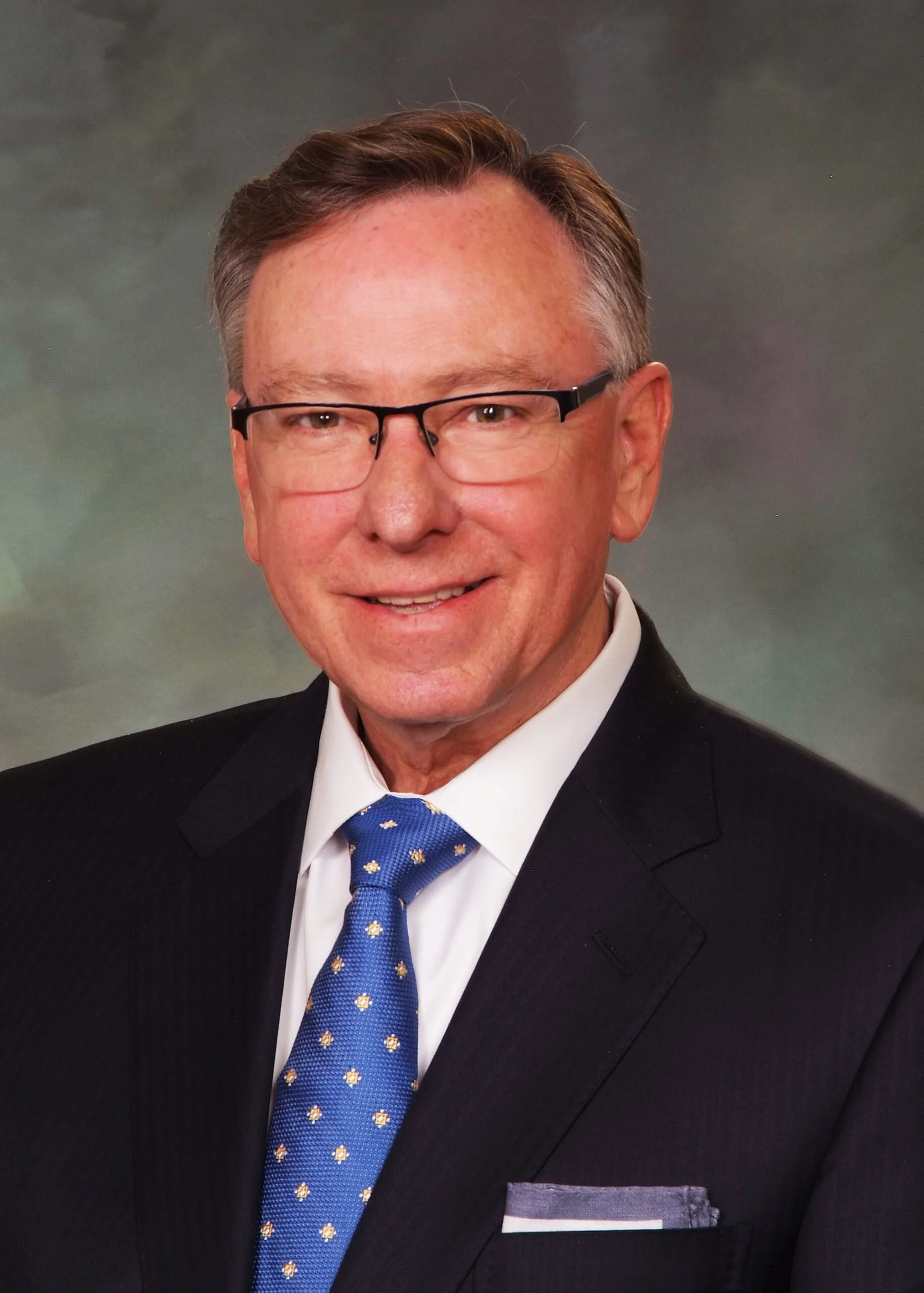 A middle-aged man with short brown hair and glasses, wearing a black suit, white shirt, and a blue tie with yellow floral patterns.