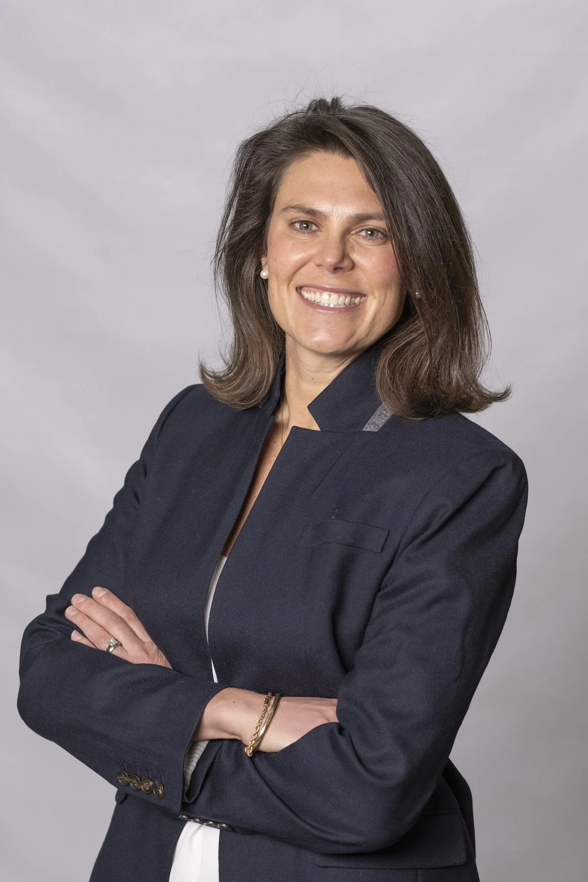 A woman with shoulder-length brown hair, wearing a dark blazer, smiling with arms crossed against a gray background.