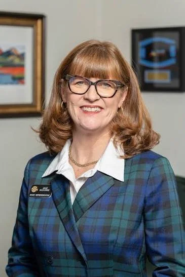 A woman with red hair, glasses, and a plaid blazer, smiling in an office setting.