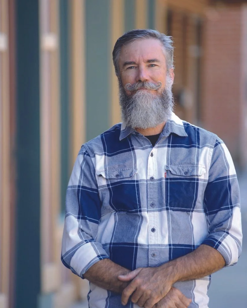A middle-aged man with a gray beard and mustache, wearing a blue and white plaid shirt, standing outside on a sidewalk during daytime, with a slight smile and arms crossed in front.
