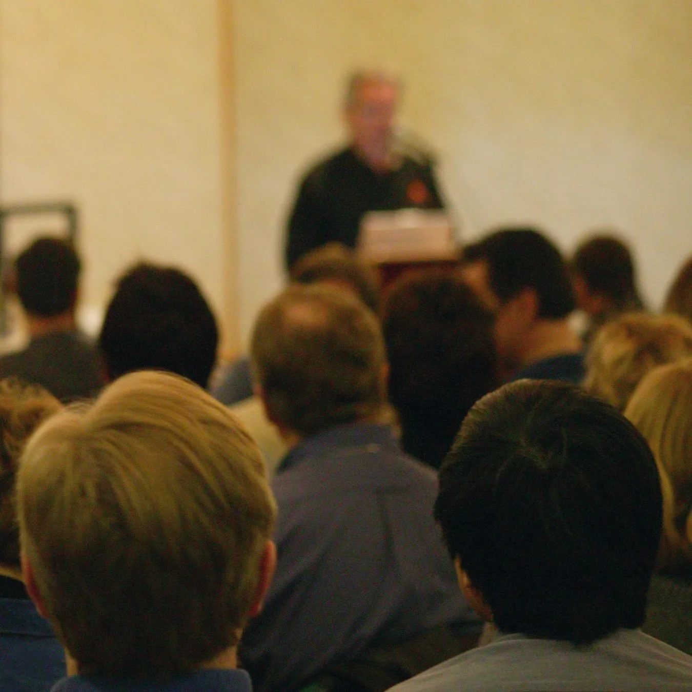 A group of people is seated and listening to Steinar Opheim at a presentation to pastors and leaders in a room with light-colored walls.