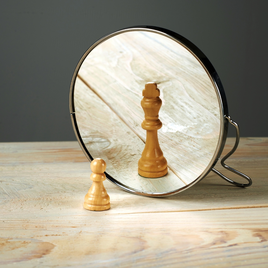 A wooden pawn chess piece is in front of a mirror that reflects its image. The pawn and mirror are placed on a light-colored wooden surface against a dark background.