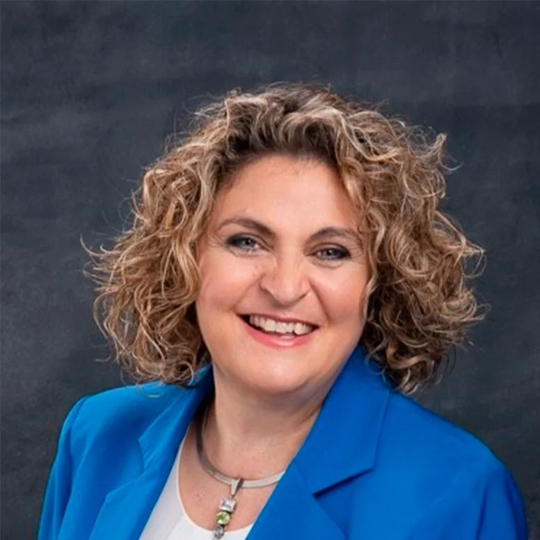 Portrait of Elizabeth Strauss, Resource Manager - Tent International. CEO of Tent SA, with curly blonde hair, wearing a blue blazer and a necklace, smiling against a dark background.