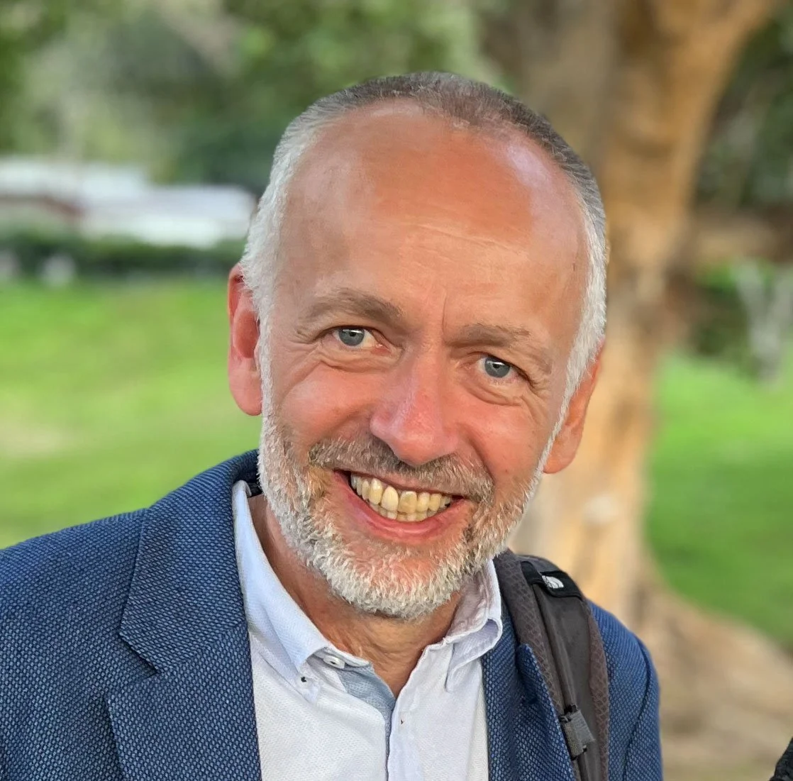 A smiling Steinar Opheim, CEO of Tent International, with gray hair and beard, wearing a blue blazer and white shirt, stands outdoors with trees and greenery in the background.