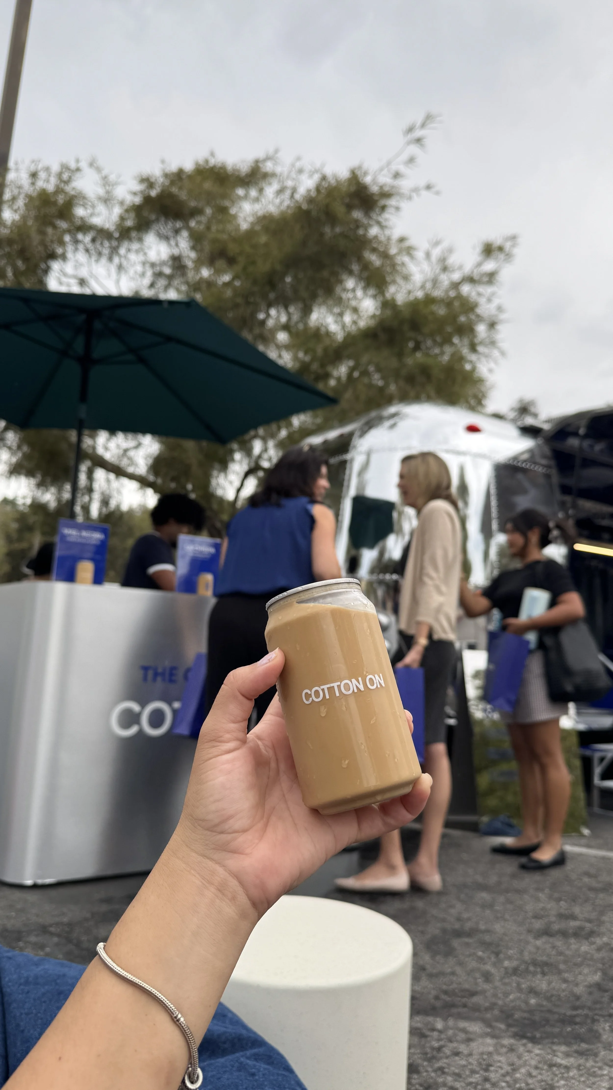 Person holding a glass of iced coffee with a crowd of people at an outdoor event in the background.