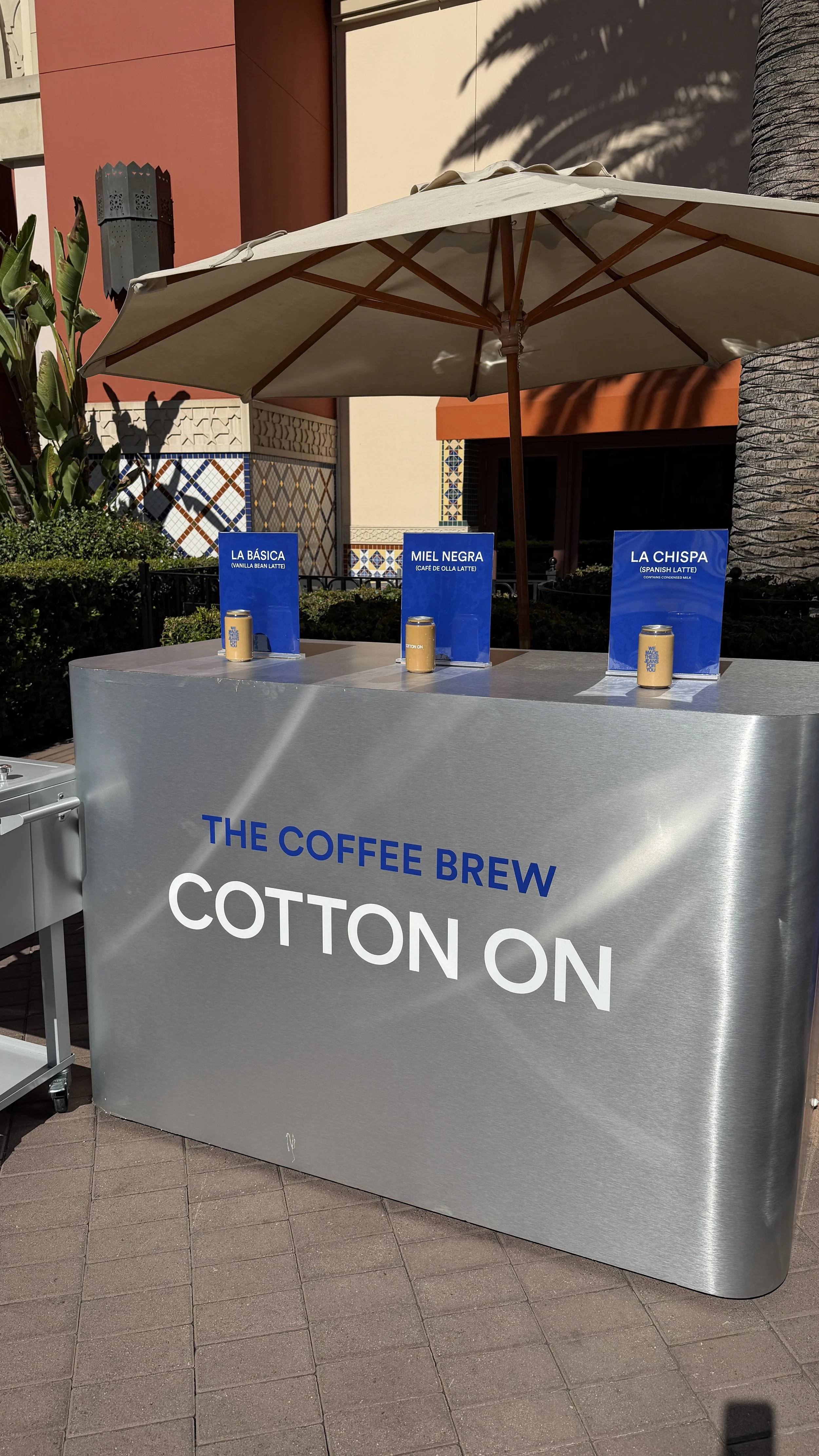A coffee stand with a silver metallic front and a large white umbrella, situated outdoors on a sunny day. The stand has blue signs displaying different coffee options, including 'La Básica,' 'Miel Negra,' and 'La Chispa,' with corresponding illustrations of canned coffee drinks.