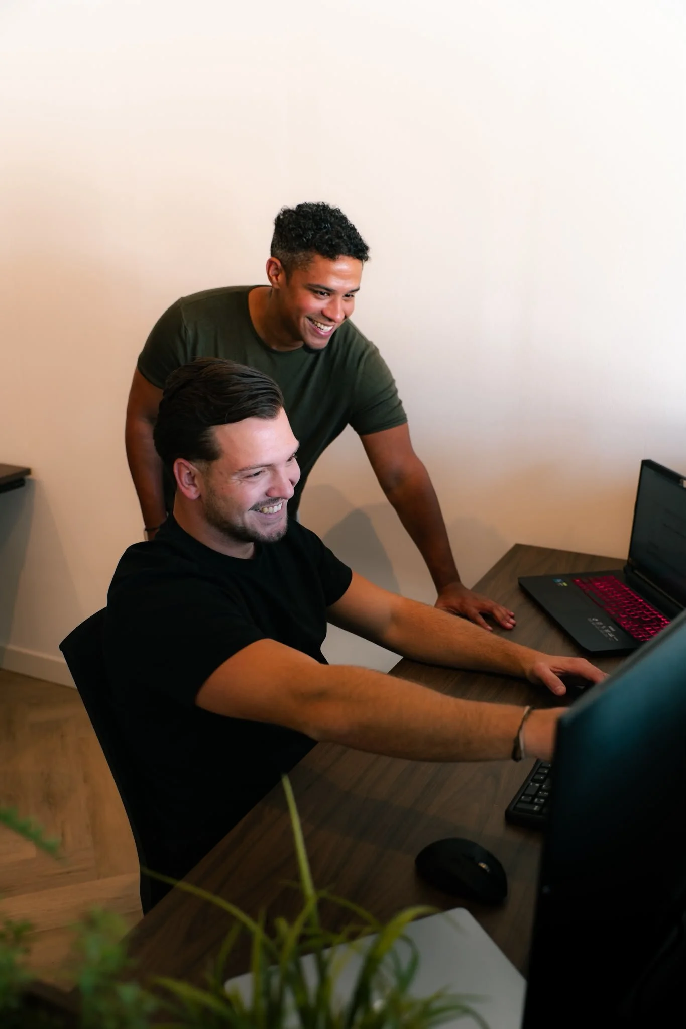 Twee mannen werken samen aan een computer. Ze lachen en kijken naar het scherm in een kantooromgeving.