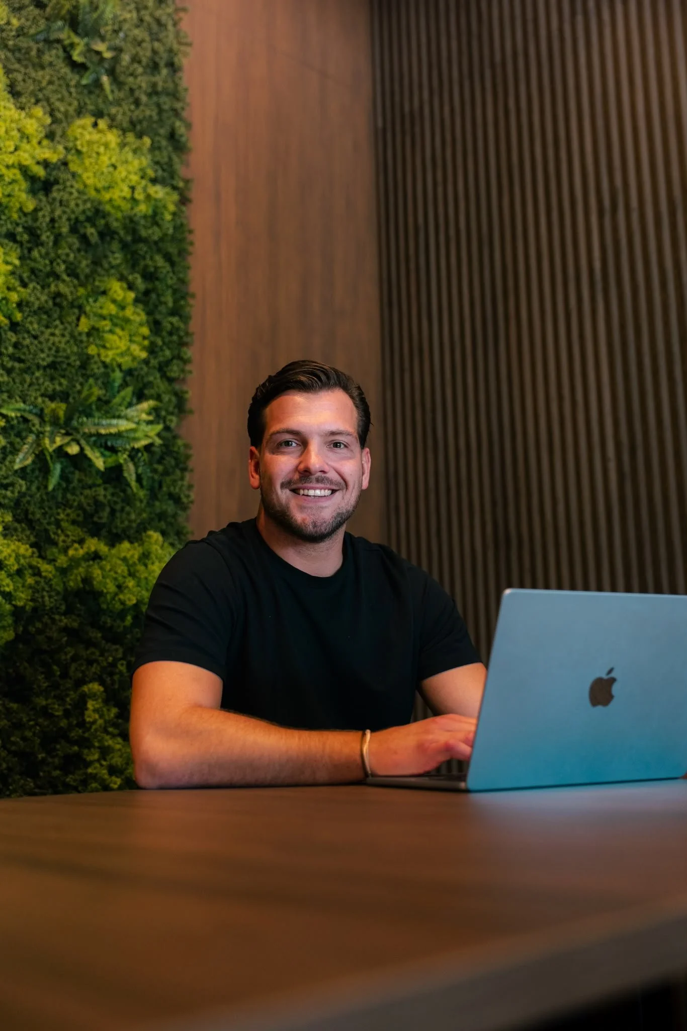 Een glimlachende man met donker haar, zittend aan een houten tafel, werkend op een zilveren MacBook laptop, binnen in een modern interieur met een groene muur en houten wandbekleding.