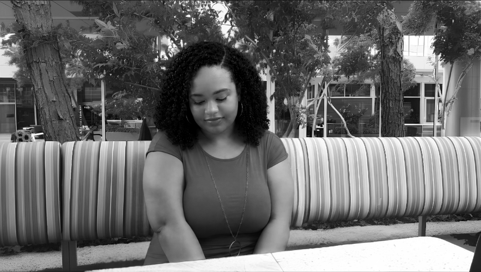 A woman with curly hair sitting on a park bench, looking down with a serious expression, outdoors with trees and buildings in the background.