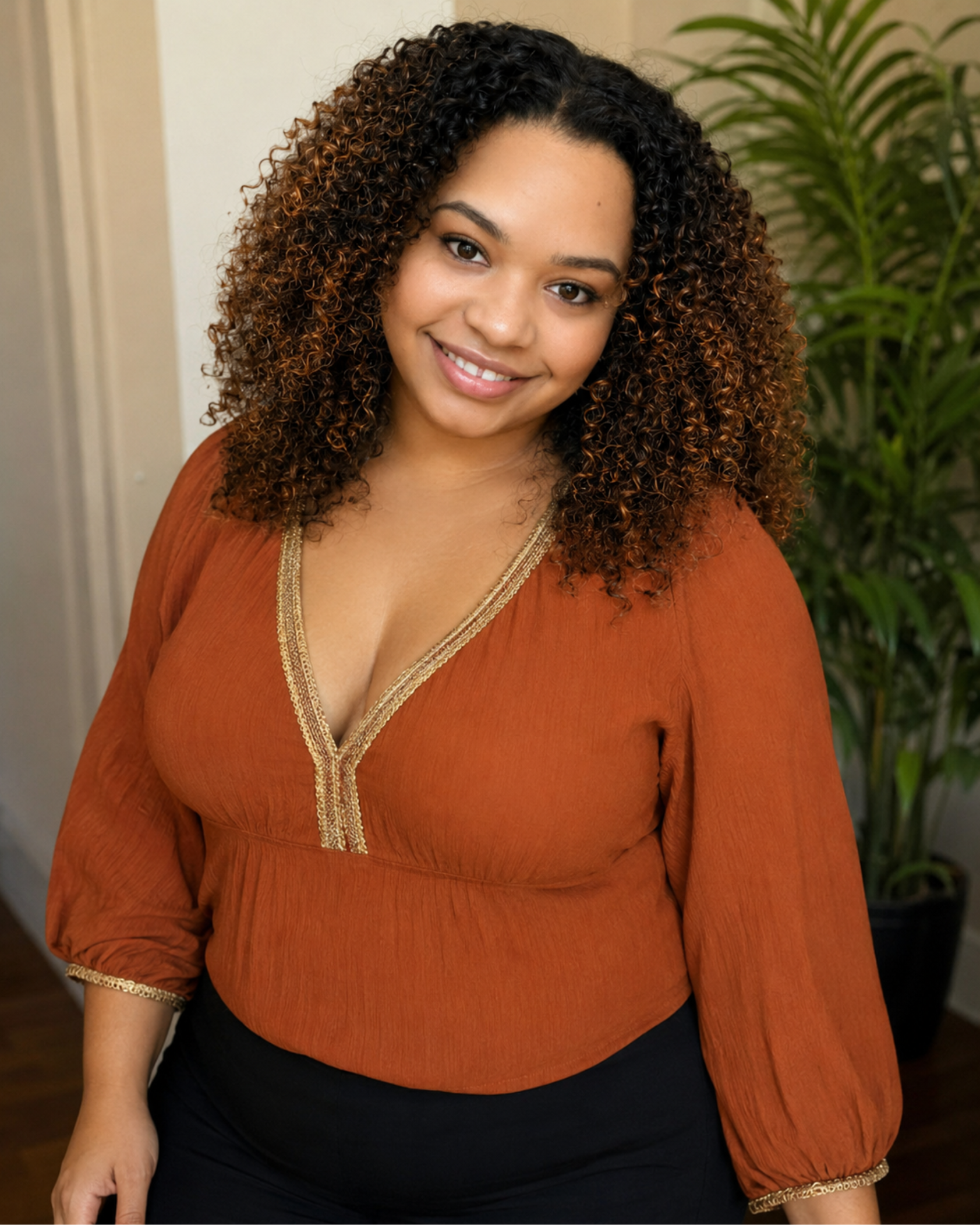 A woman with curly hair wearing an orange top with gold trim, standing indoors by a green plant, smiling at the camera.