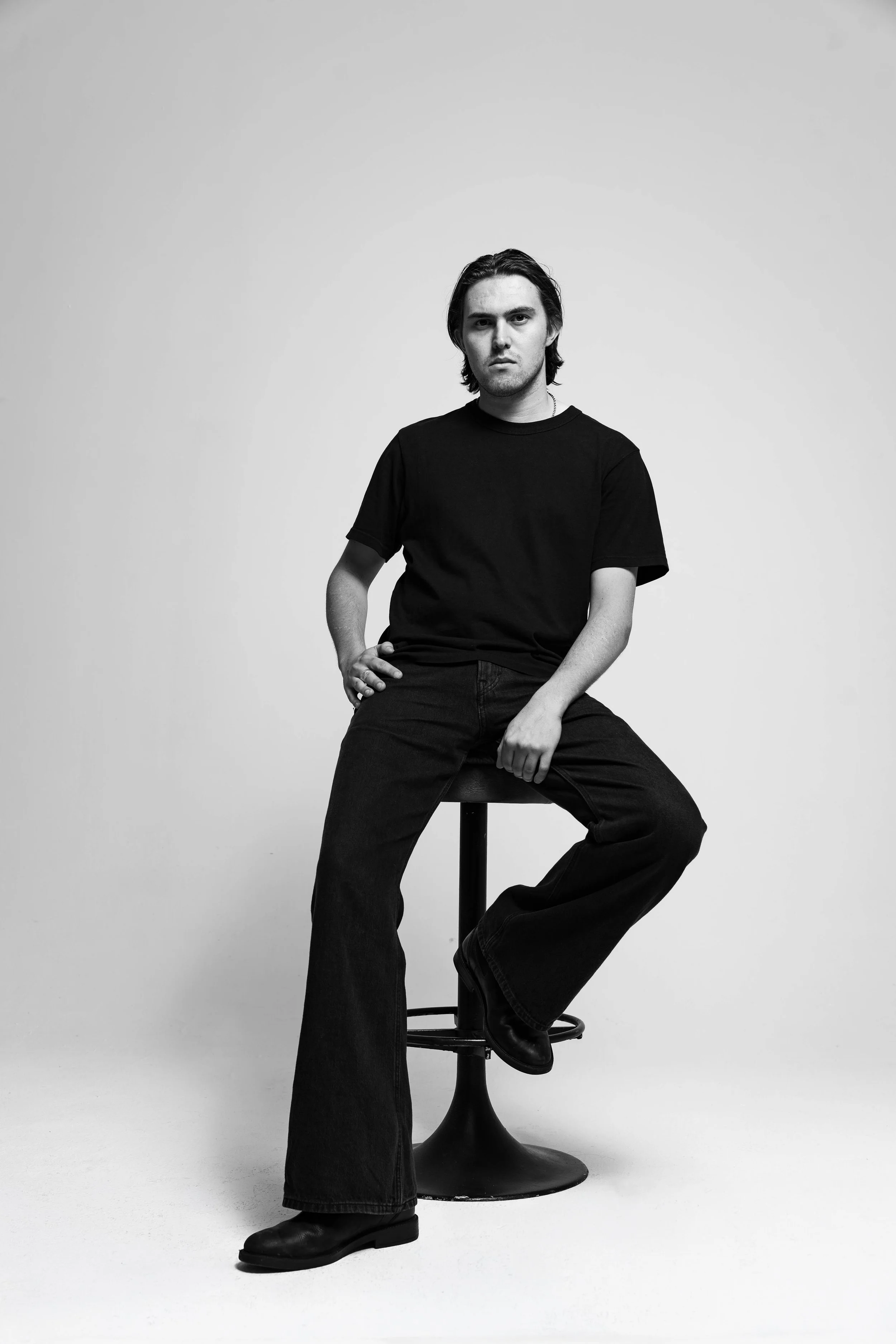 A young man with medium-length dark hair and a serious expression, sitting on a stool with one hand on his hip in a black t-shirt, black jeans, and black shoes, in a minimalistic studio setting with a plain light background.