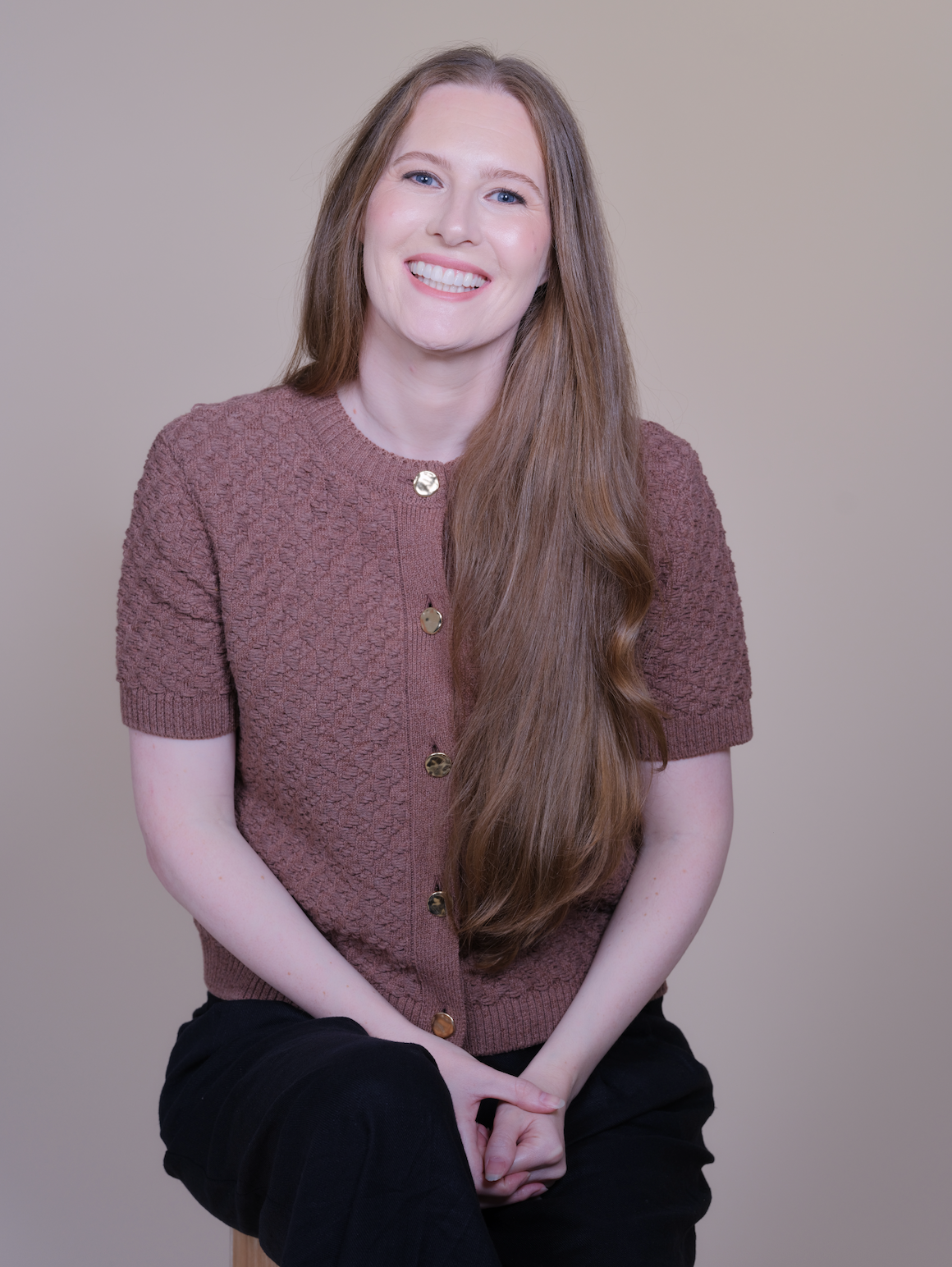 A woman with long hair, smiling, wearing a brown short-sleeved cardigan with gold buttons, sitting against a plain neutral-colored background.
