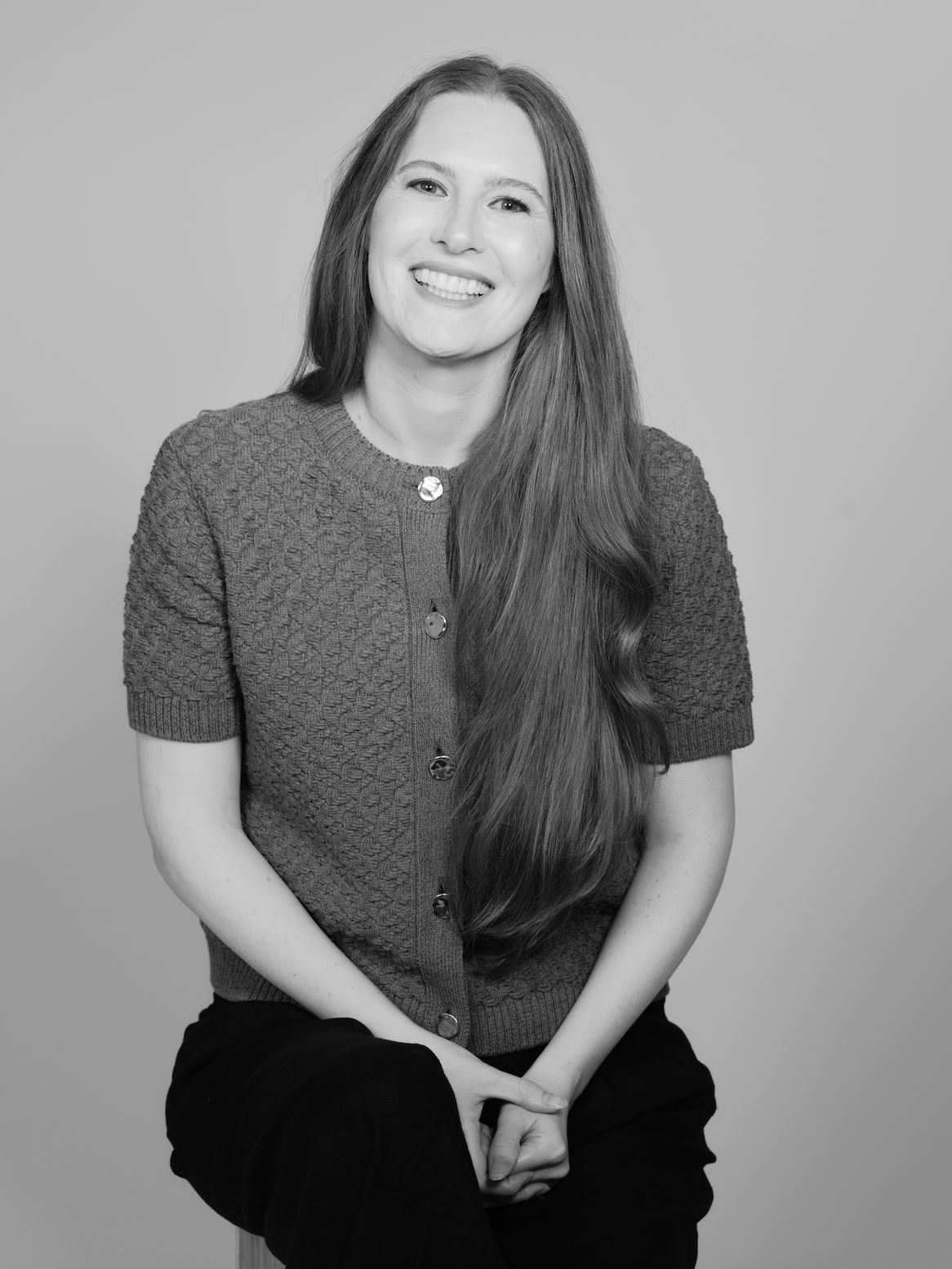 Black and white portrait of a smiling woman with long, straight hair, wearing a textured short-sleeve cardigan and dark pants, sitting against a plain background.