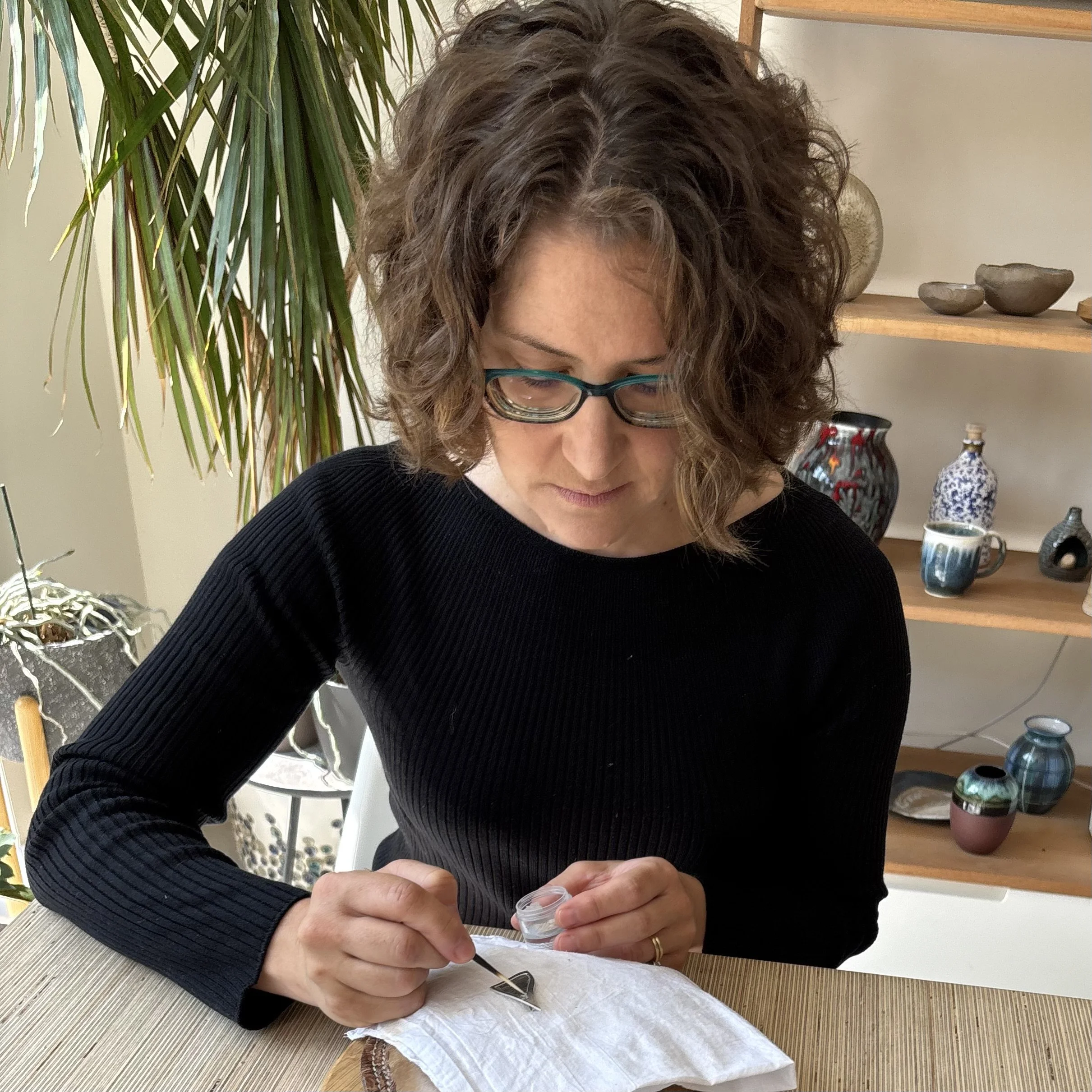 Woman with curly hair and glasses examining small objects on a white cloth, in a room with shelves displaying pottery.