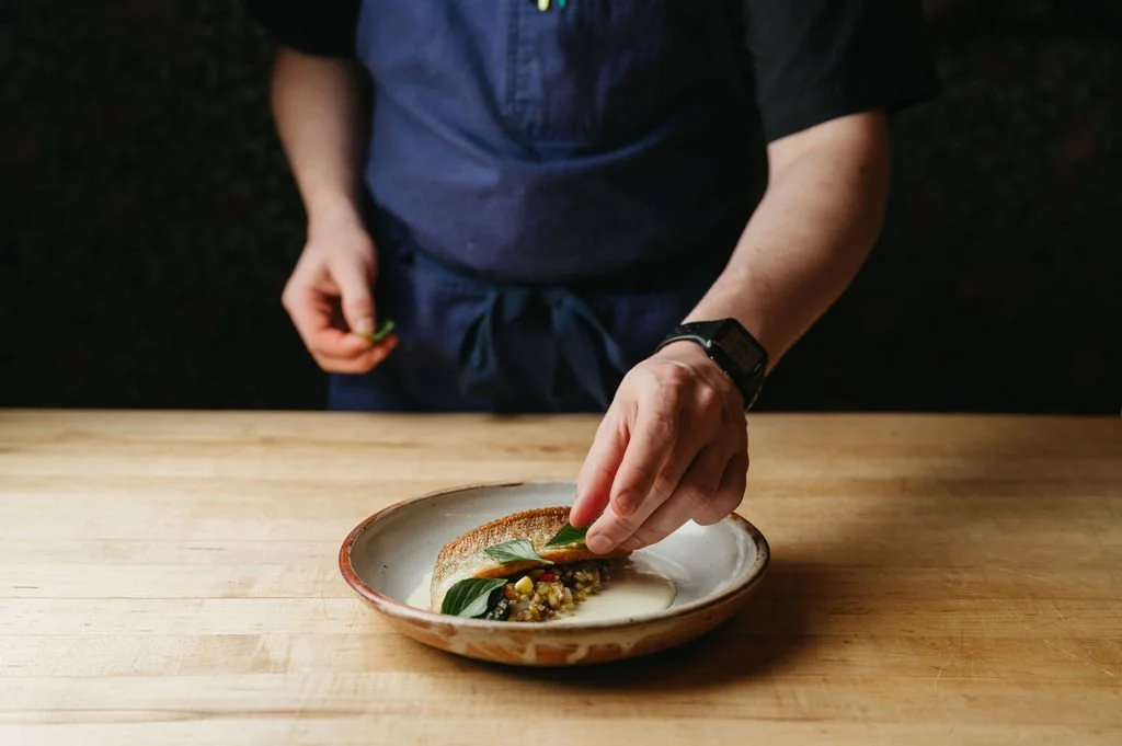 Person placing a green herb on a plated dish with a piece of toasted bread and a creamy sauce with vegetables, on a wooden table.