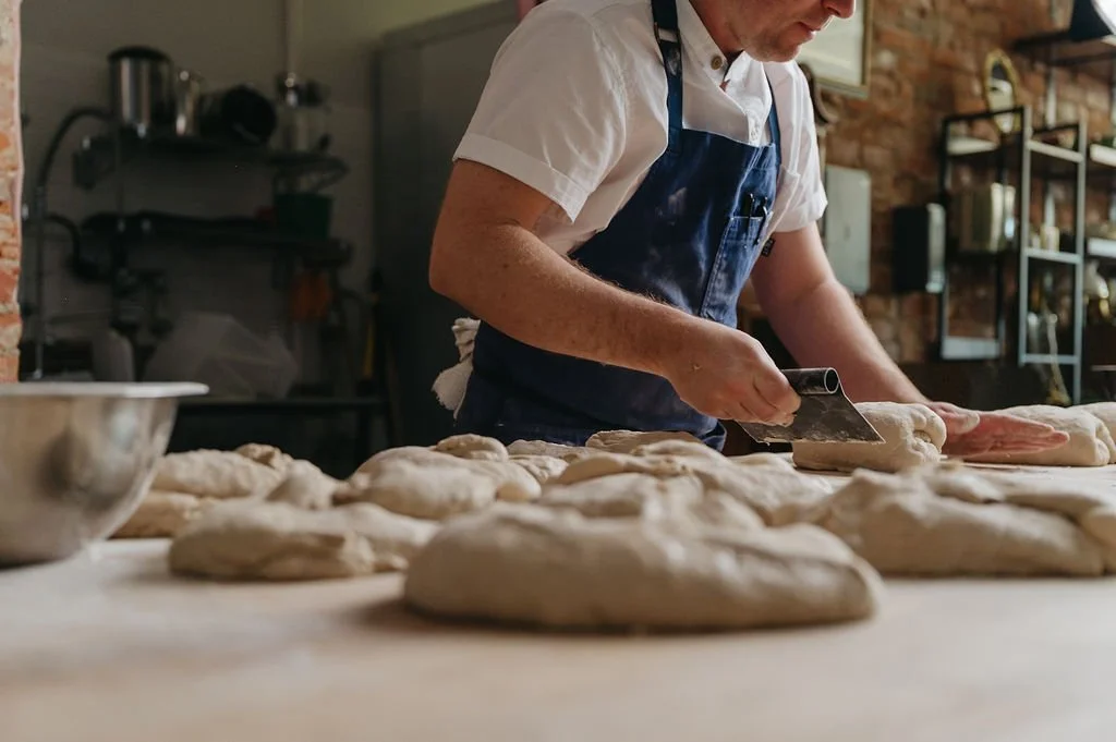 A person in a white shirt and blue apron is slicing dough on a wooden surface in a bakery kitchen. Several dough pieces are laid out on the table, and there are shelves with baking supplies in the background.
