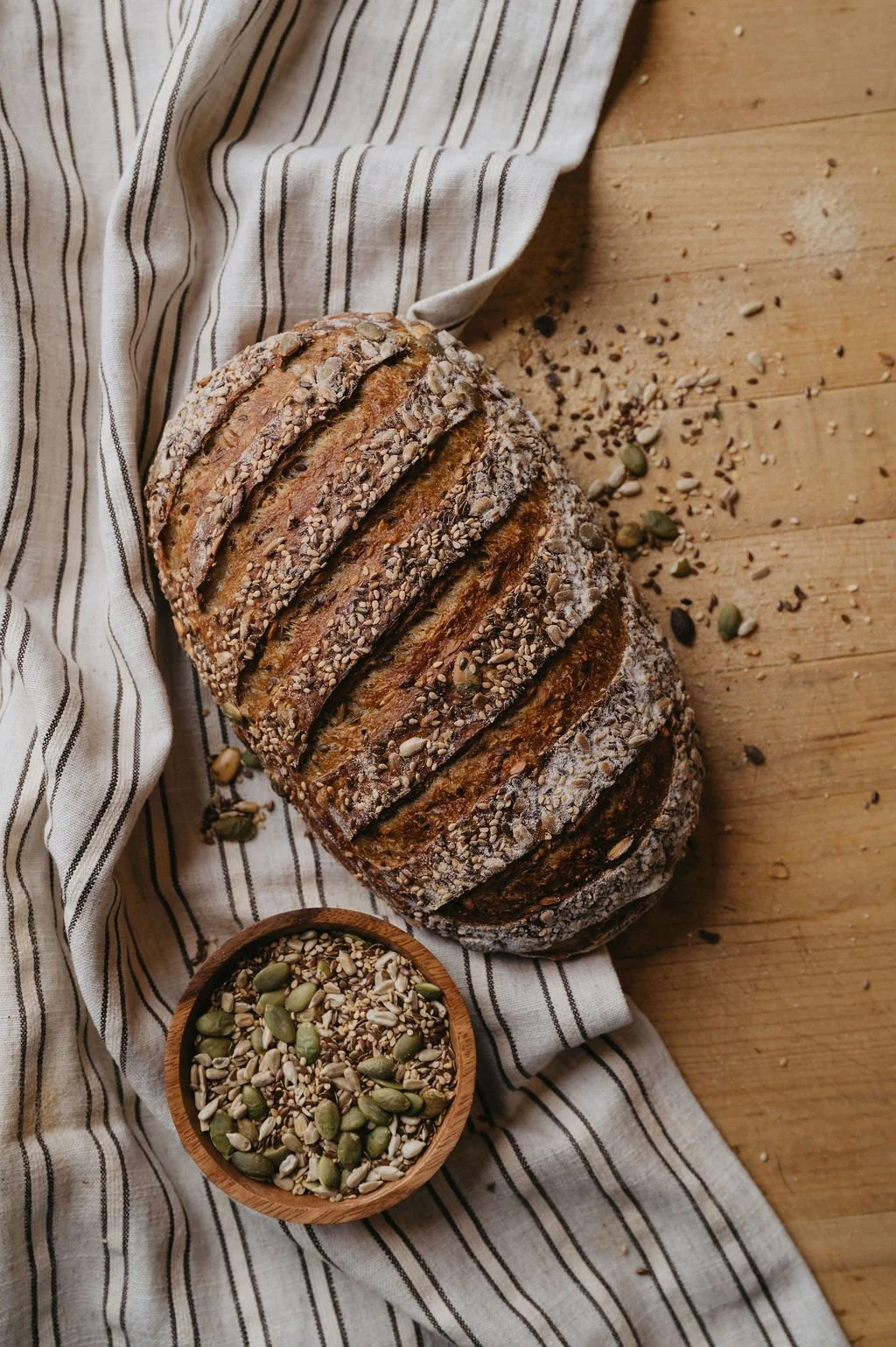 Sliced multigrain bread with seeds on a striped cloth and wooden surface, with a small bowl of mixed seeds nearby.