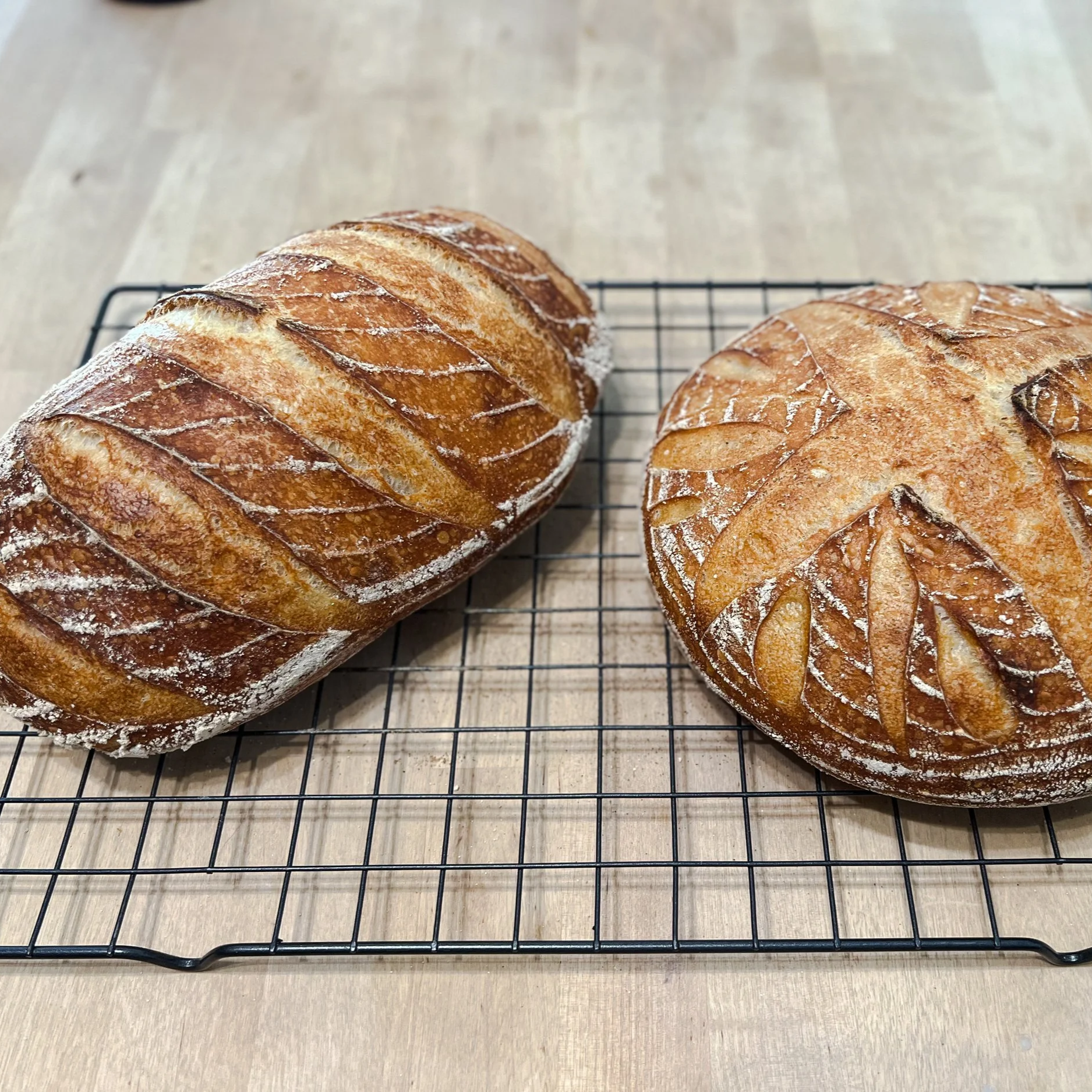 Two freshly baked baguette loaves with scored tops resting on a cooling rack on a light-colored wooden surface.