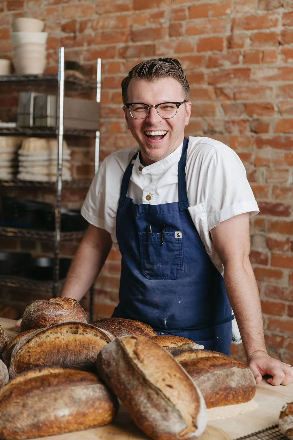 A smiling man wearing glasses and a white shirt with a navy apron, standing behind a table with various loaves of bread, in a bakery with a brick wall background.