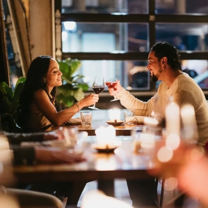 A couple enjoying drinks and toasting in a warmly lit restaurant with candles and large windows.