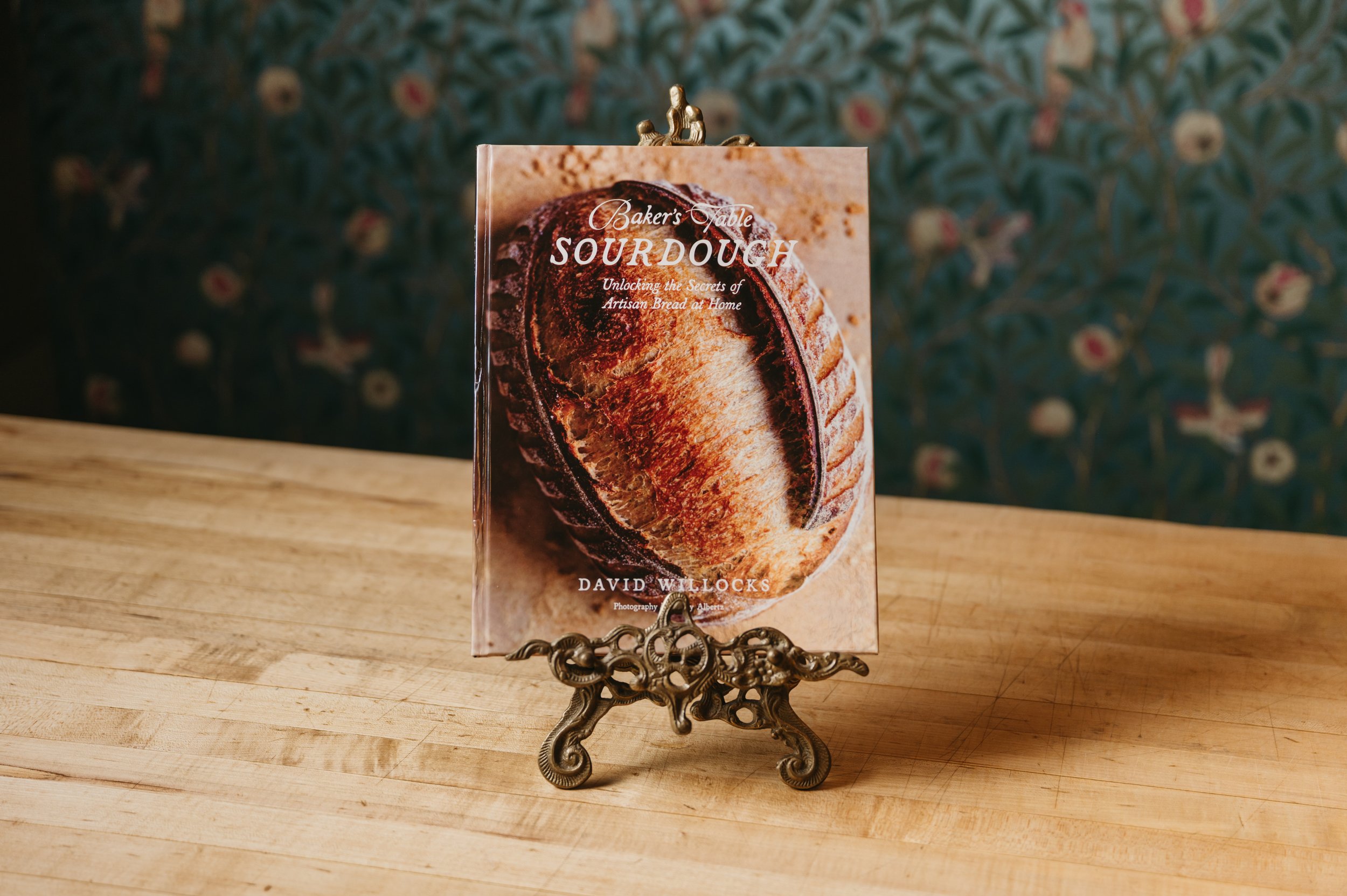 Book titled 'Baker's Table Sour Dough' standing on an ornate metal stand on a wooden surface, with a blurred floral wallpaper background.