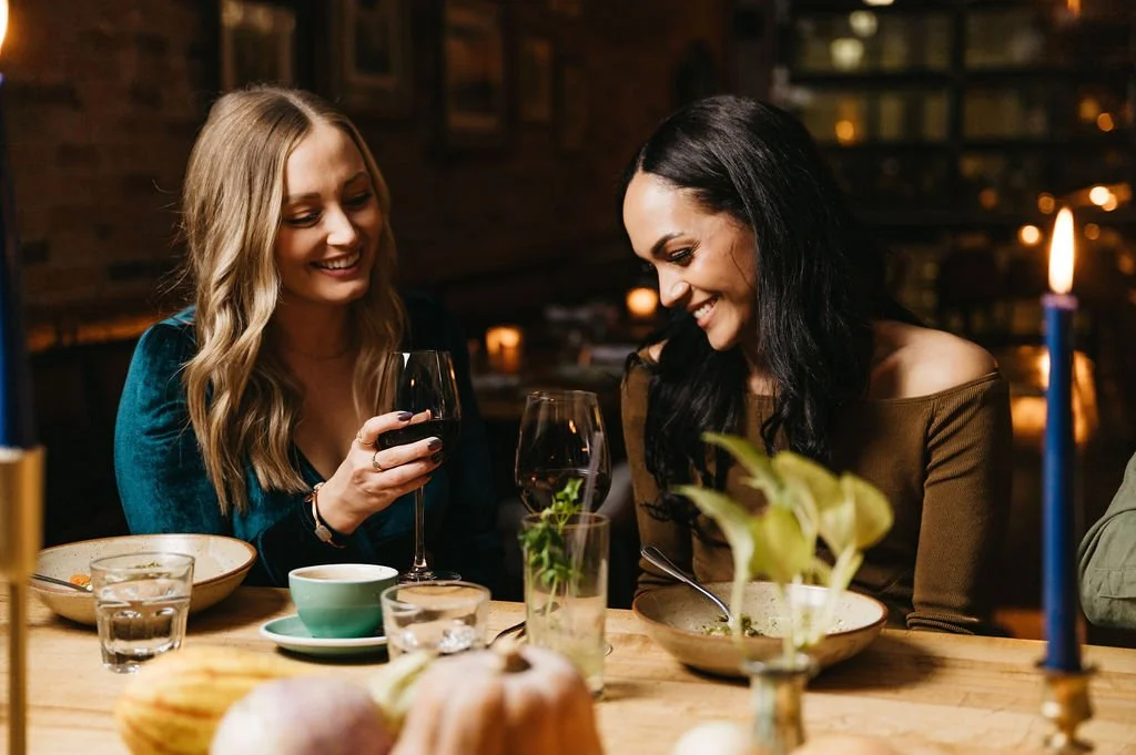 Two women enjoying a dinner at a cozy restaurant, one holding a glass of red wine, surrounded by plates, glasses, and candles on a wooden table.