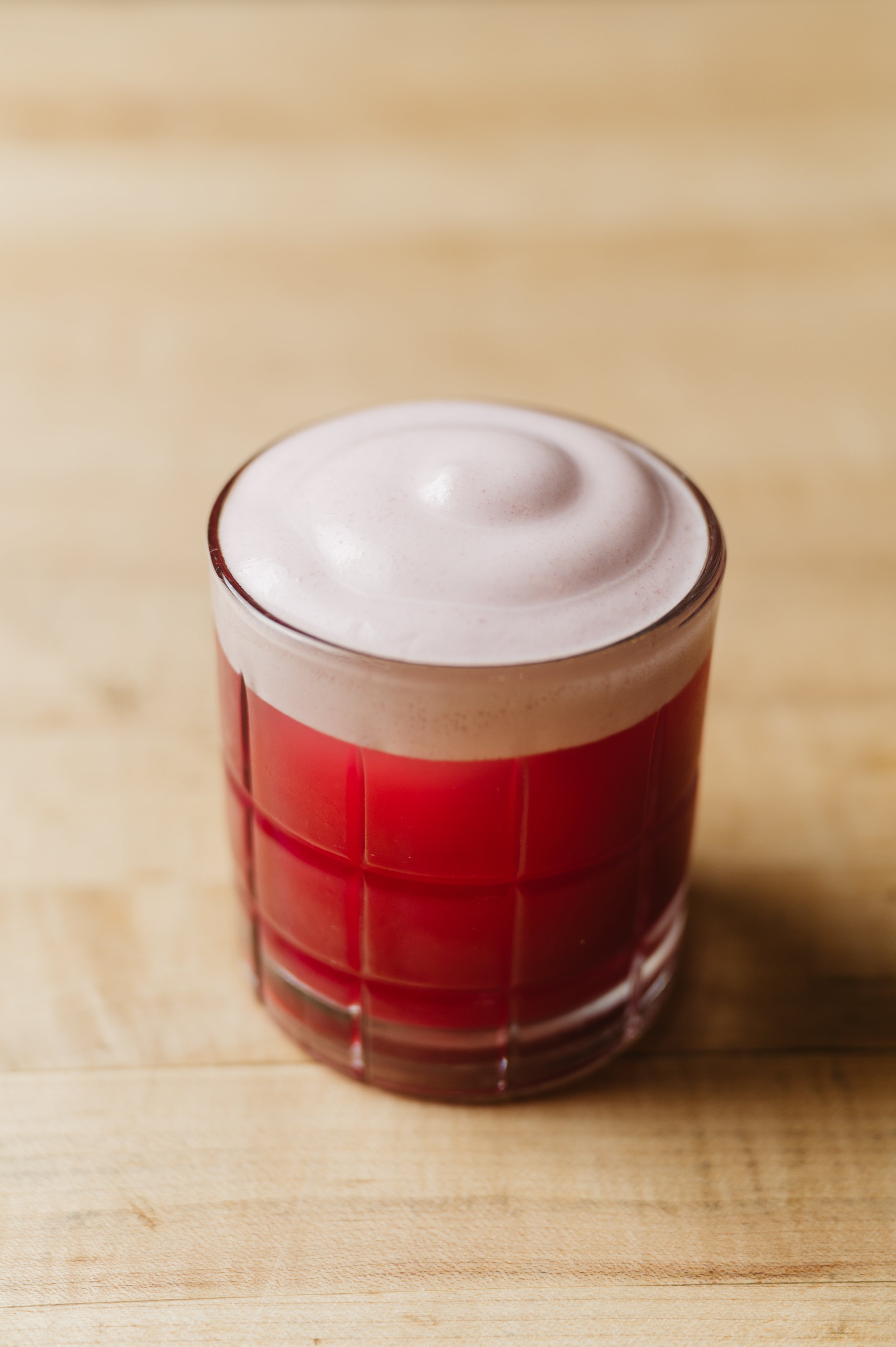 A close-up of a glass of red liquid with foam on top, placed on a wooden surface.